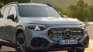 Front view of a silver Mercedes-Benz SUV with illuminated grille and headlights on a road with trees in the background.