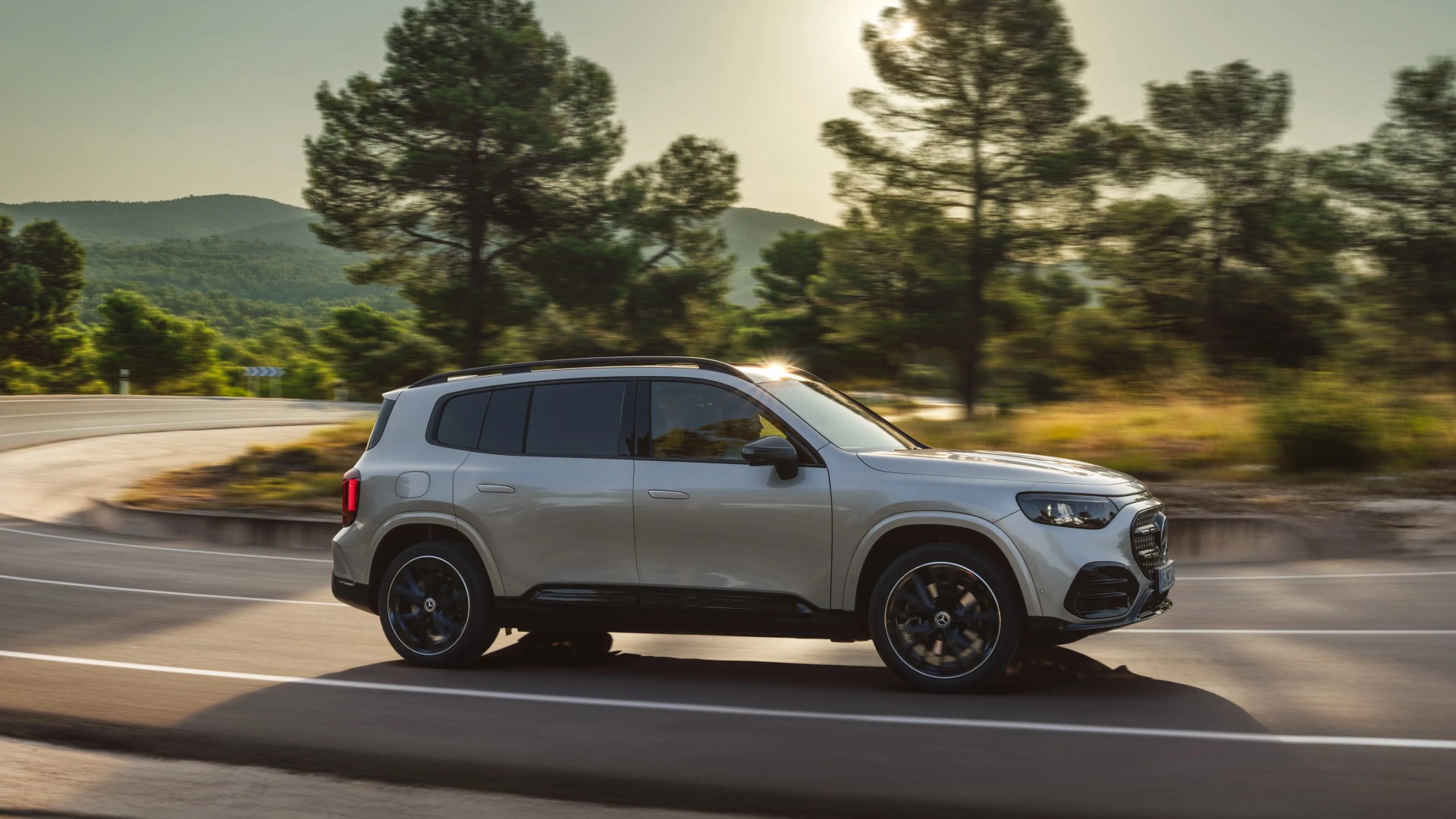 Silver Mercedes-Benz SUV driving on a curved road with trees and hills in the background.