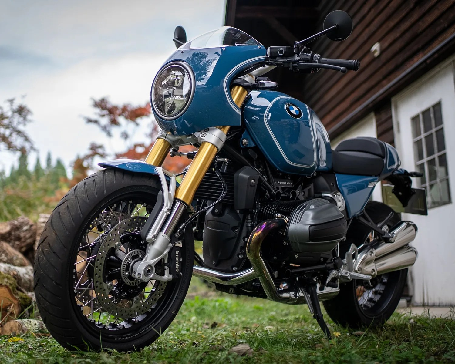 Blue BMW motorcycle with gold front forks and round headlight parked on grass near a wooden building.