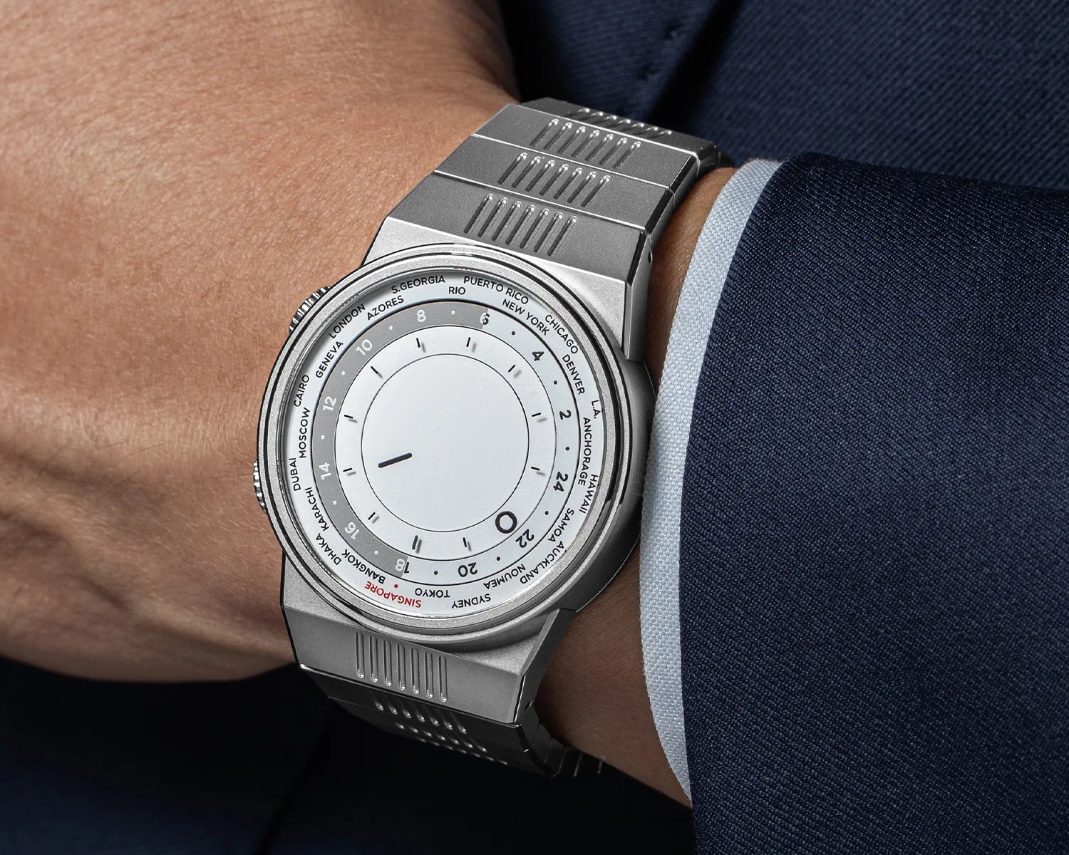 Silver metal wristwatch with a white dial displaying world cities and a single black hand, worn on a wrist with a navy suit.
