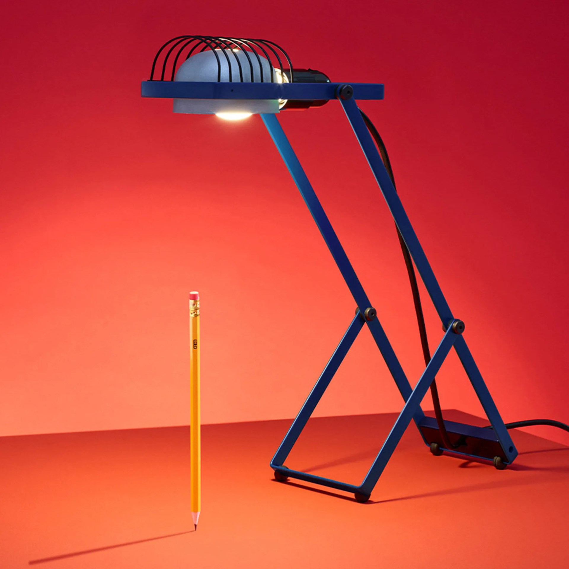 Blue adjustable desk lamp with a protective cage over the bulb illuminating a yellow pencil standing upright on an orange surface.