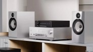 Silver stereo amplifier with black volume knob and black audio device on top, flanked by two silver speakers on a white shelf.