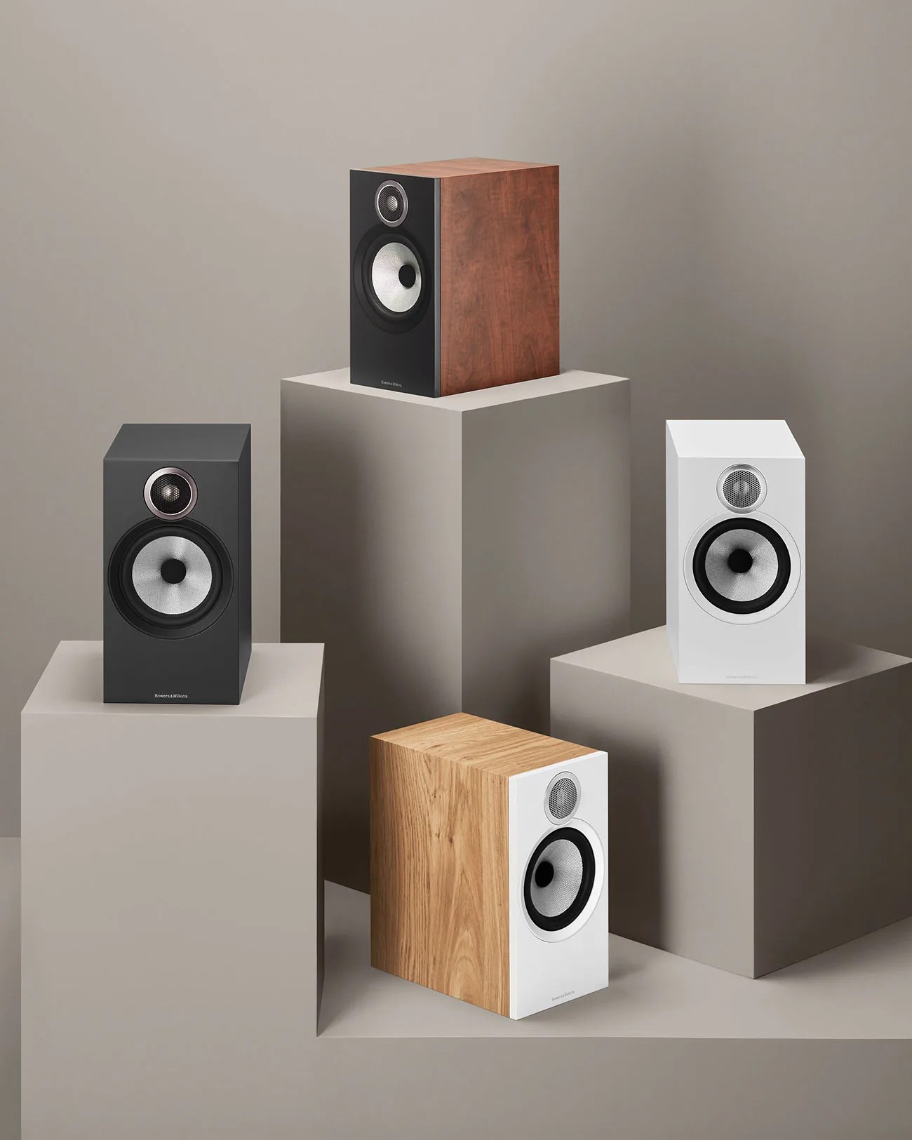 Four bookshelf speakers in different finishes—black, walnut wood, white, and oak wood—arranged on beige geometric pedestals.
