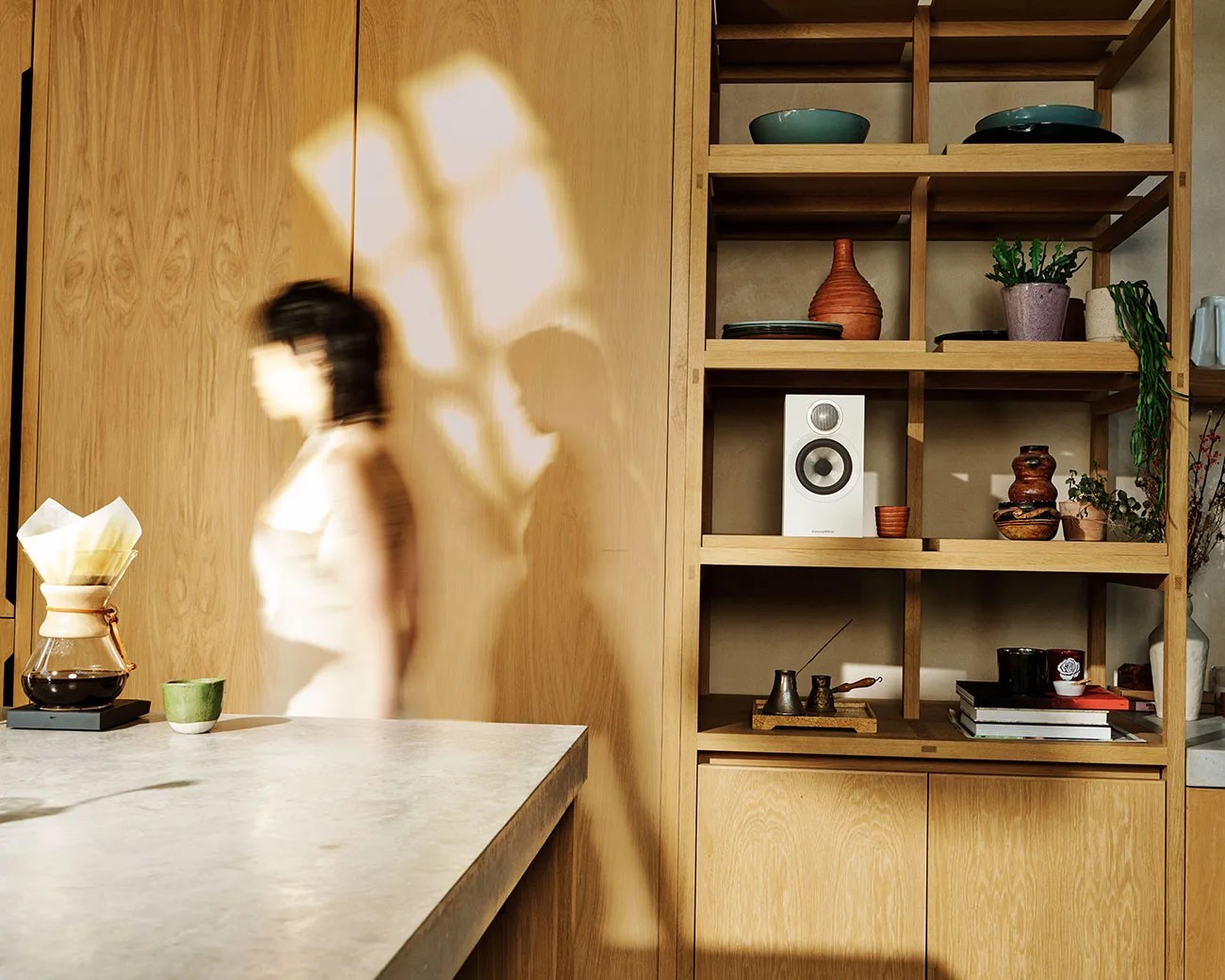 Blurred person walking past a wooden shelf with pottery, plants, books, and a white speaker in a sunlit room.