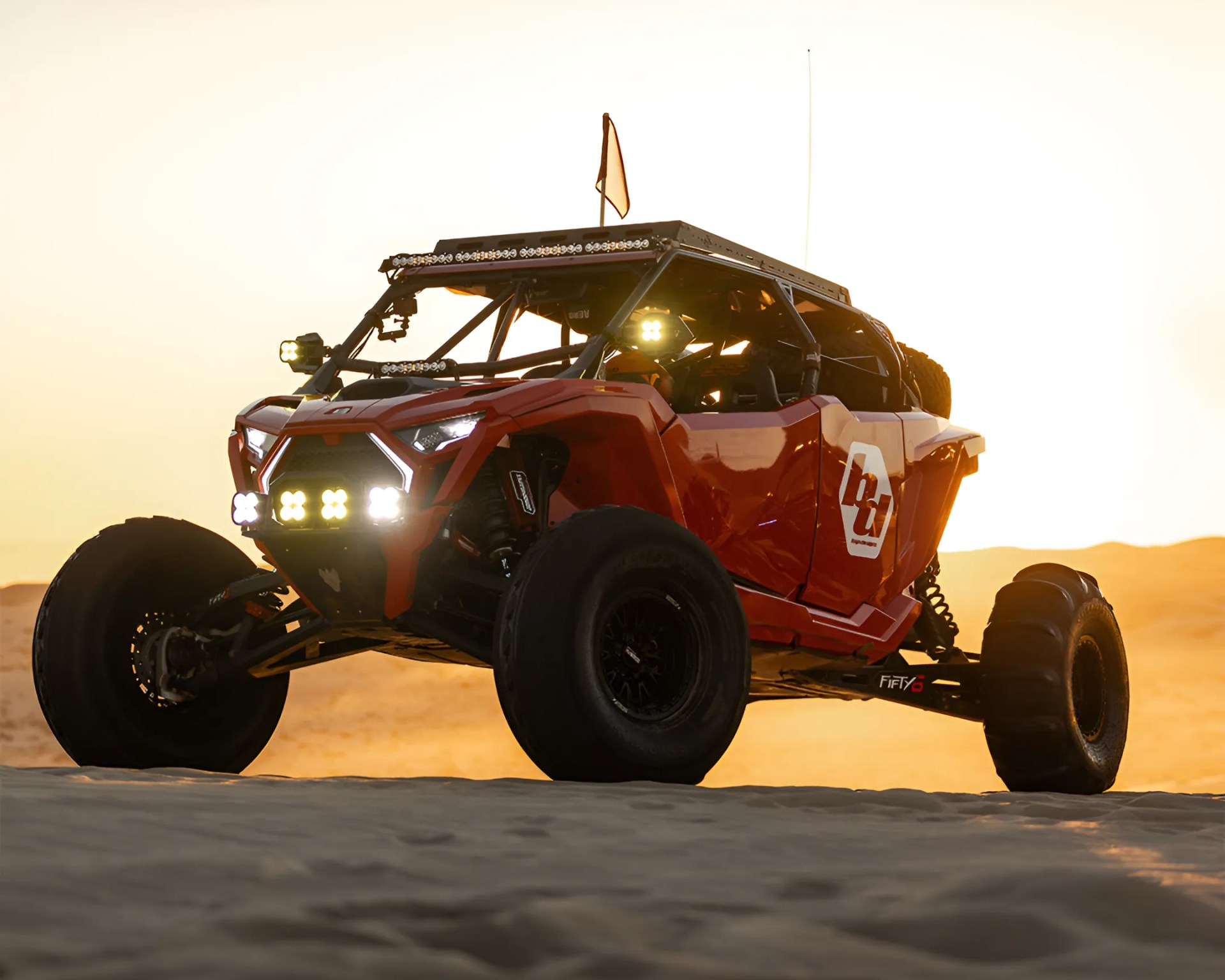 Red off-road dune buggy with multiple LED lights and large tires on sand at sunset.