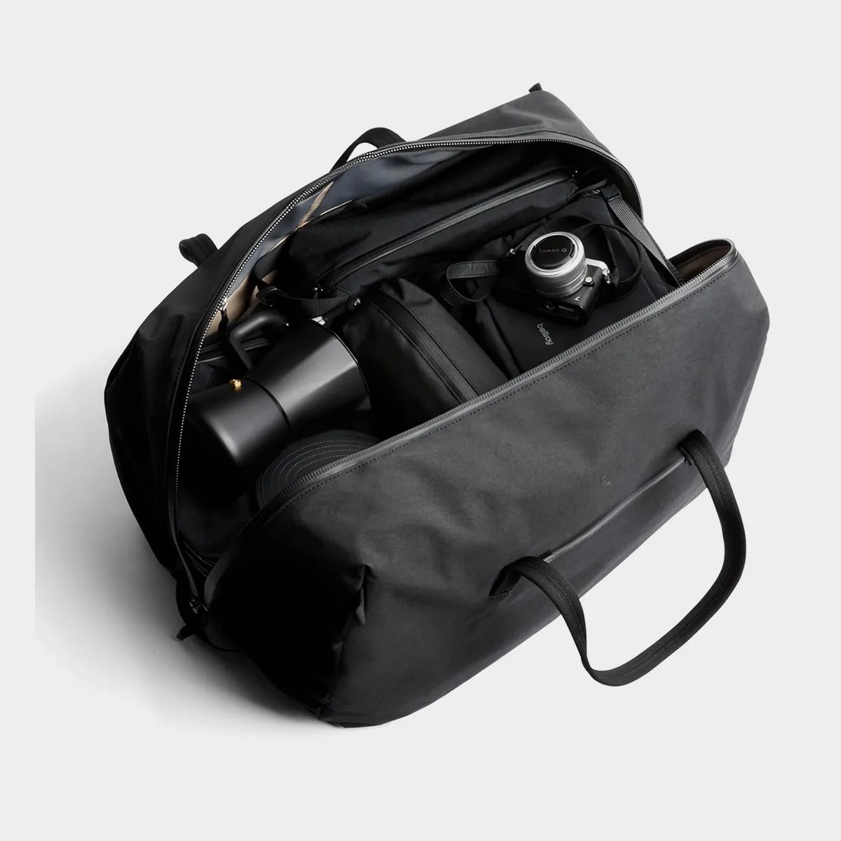Black duffel bag with top zipper open, containing a camera, coffee maker, and other black travel accessories.