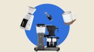 Black and silver Moccamaster coffee maker with glass carafe, white Baratza coffee grinder, black French press, and white ceramic mug with brown base.