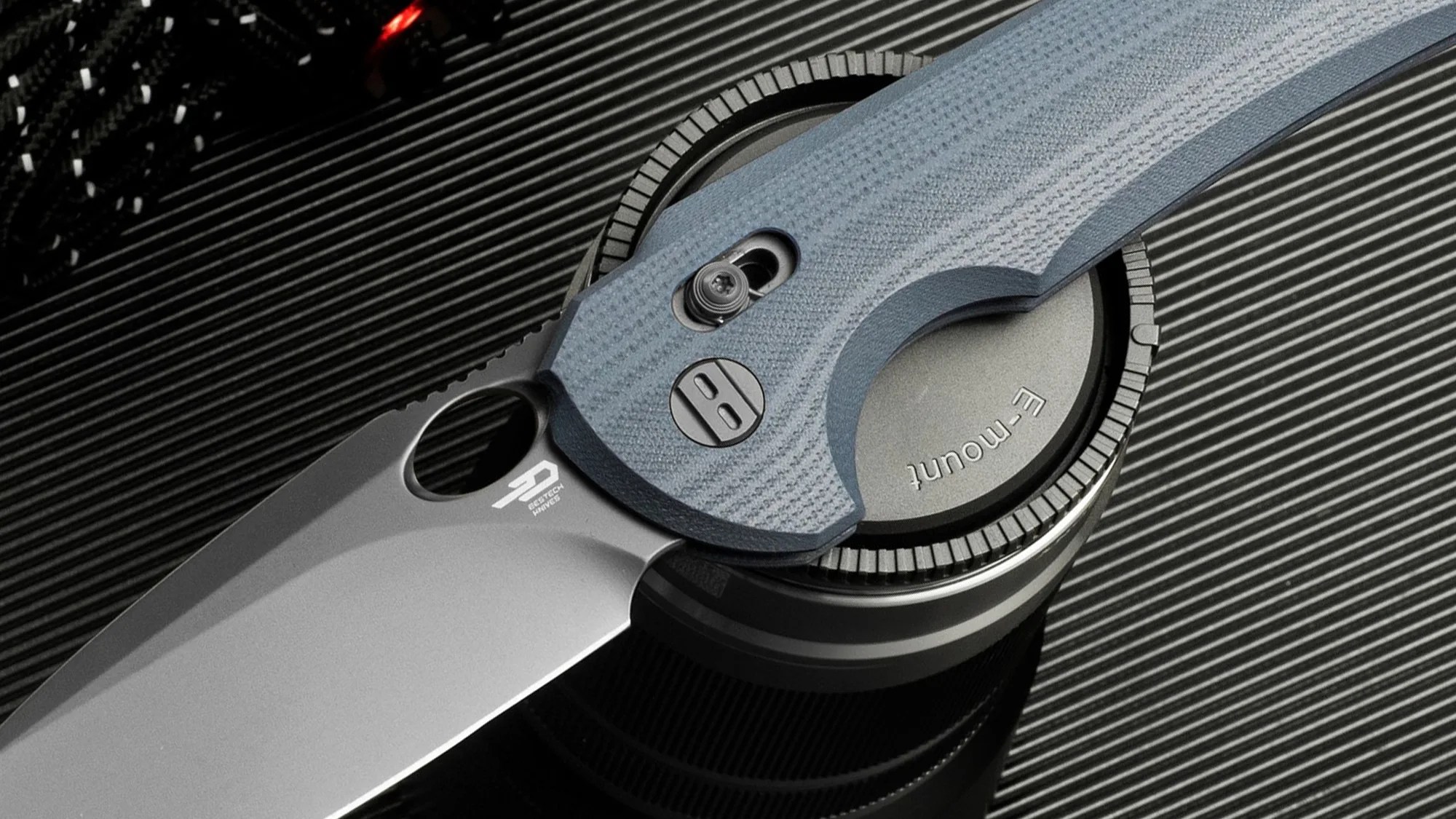 Gray folding knife with textured blue handle resting on a camera lens cap labeled "E-mount.