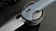 Gray folding knife with textured blue handle resting on a camera lens cap labeled "E-mount.