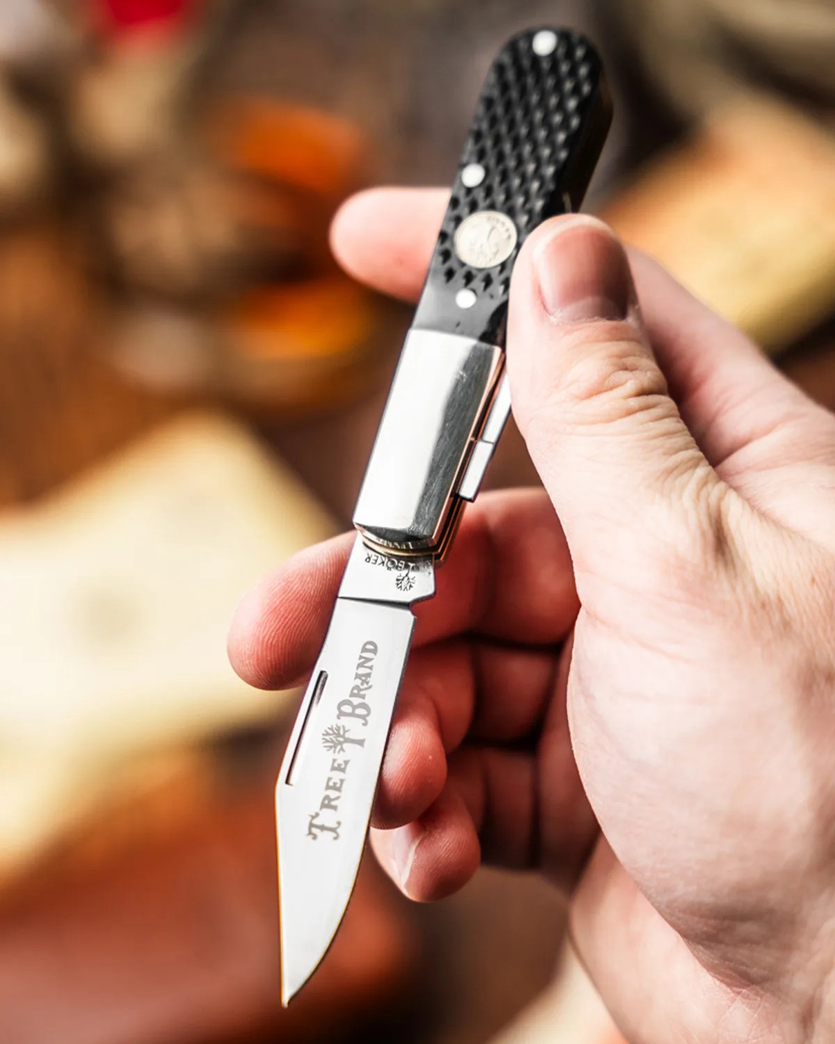 Hand holding a folding pocket knife with a black textured handle and "Tree Brand" engraved on the blade.