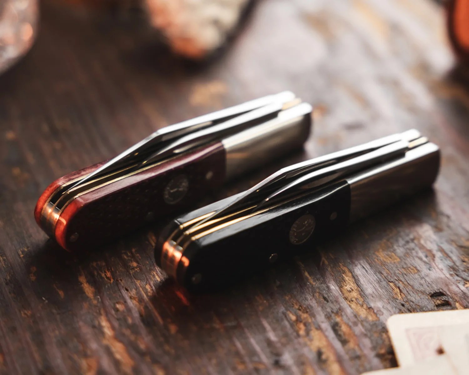 Two closed pocket knives with metal clips, one with a dark red handle and the other with a black handle, on a wooden surface.