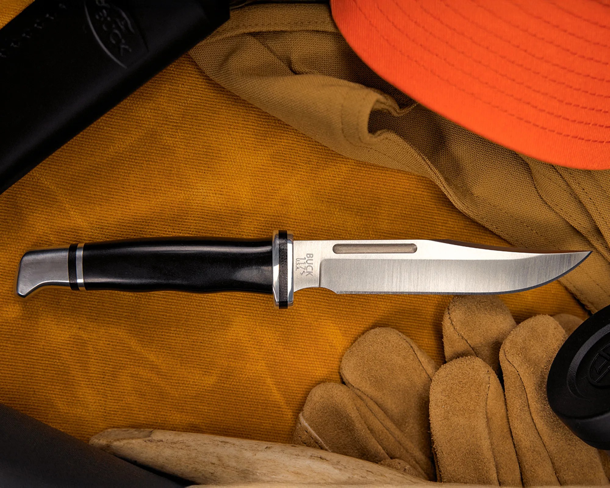 Buck 119 fixed blade knife with black handle on a mustard fabric background with tan gloves and an orange hat.