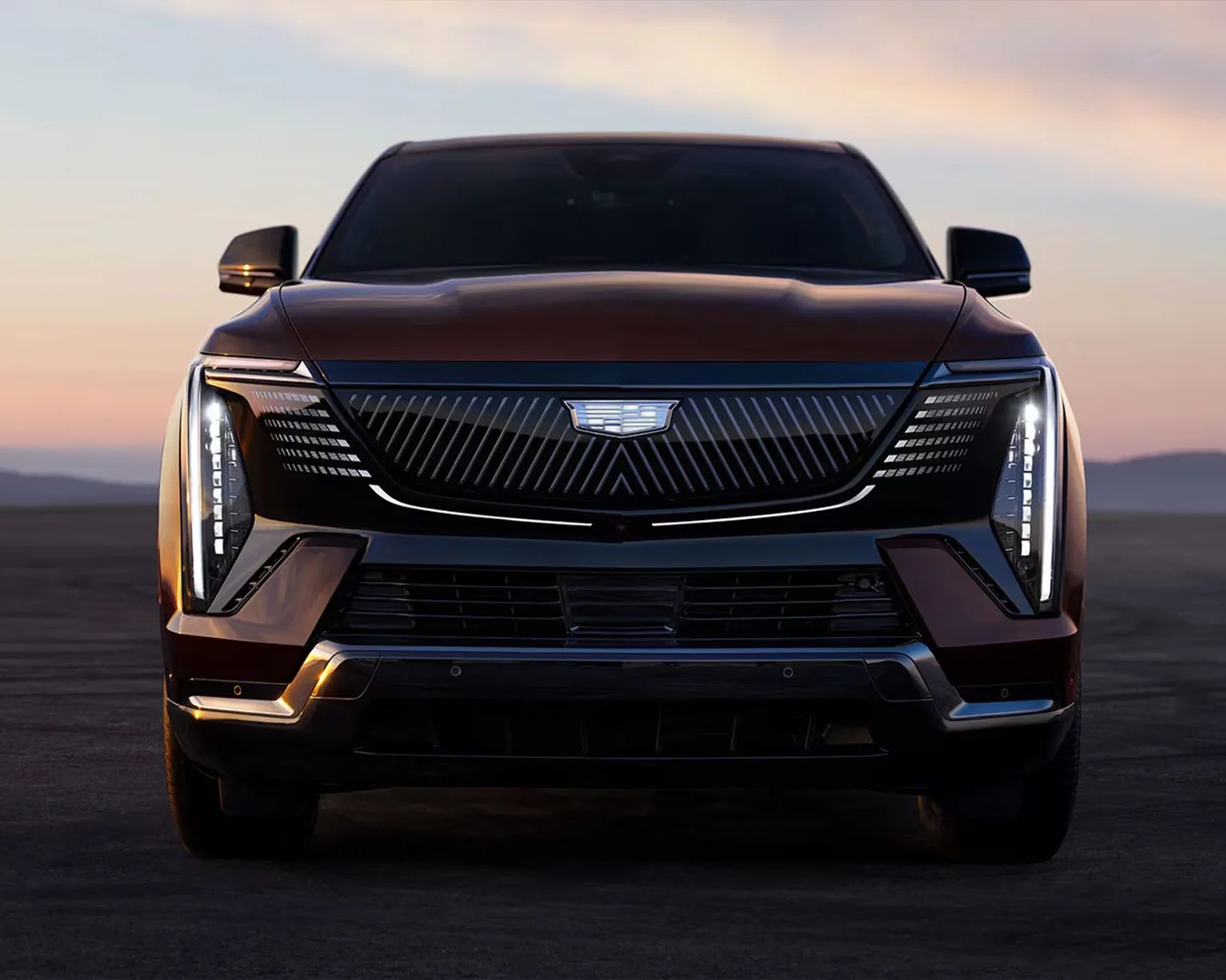 Front view of a dark-colored Cadillac SUV with illuminated vertical LED headlights at dusk.