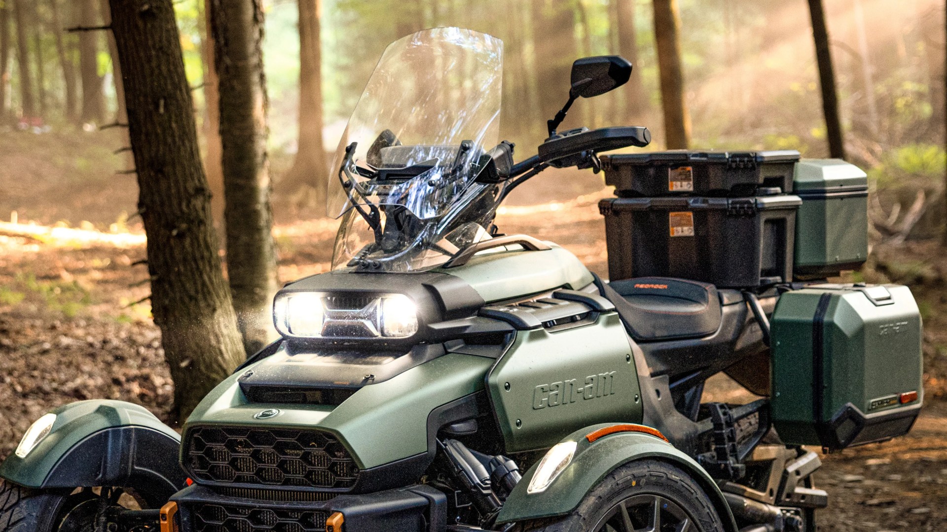 Green Can-Am three-wheeled motorcycle with windshield and multiple storage boxes in a forest setting.