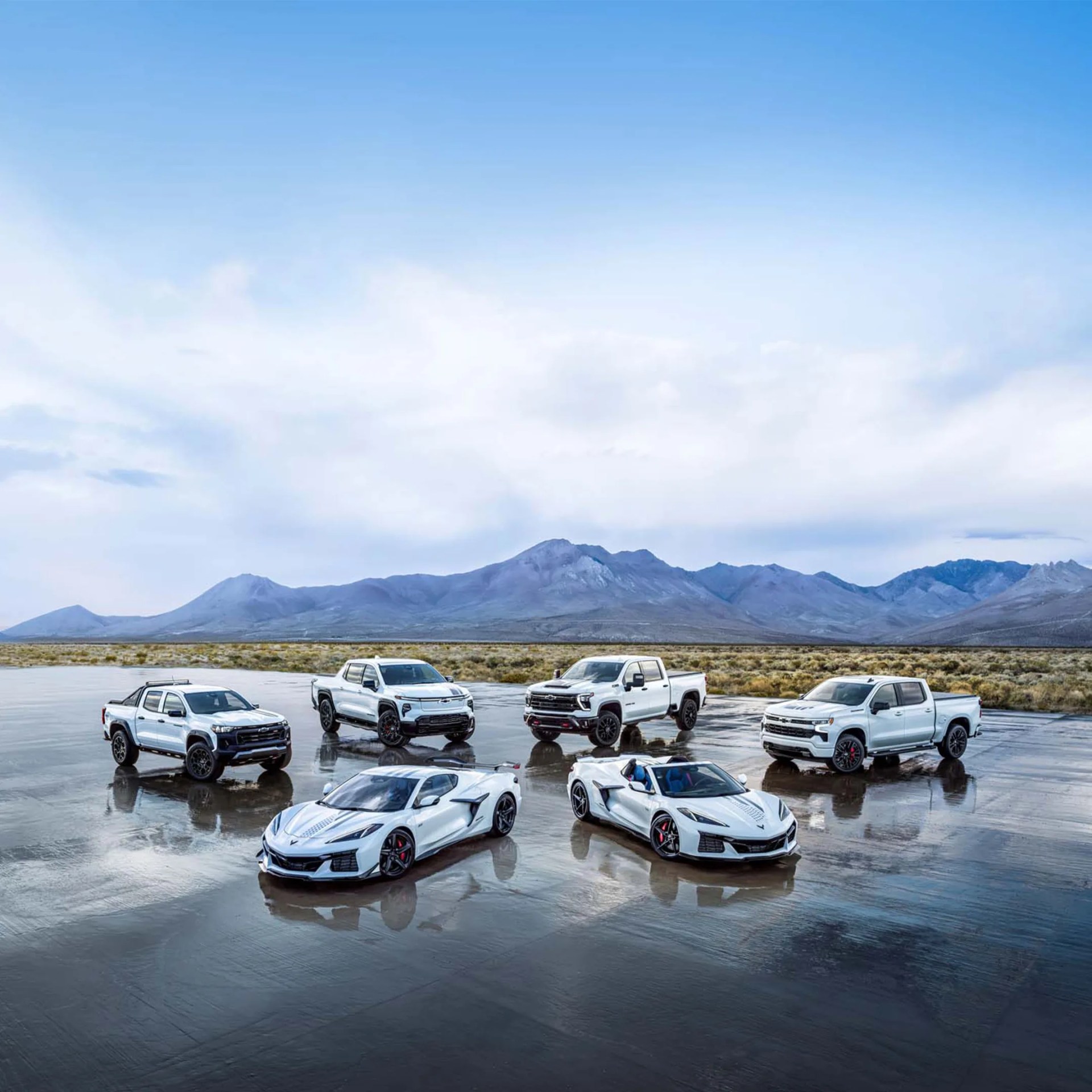 Five white Chevrolet vehicles, including two sports cars and three pickup trucks, parked on a wet reflective surface with mountains in the background.