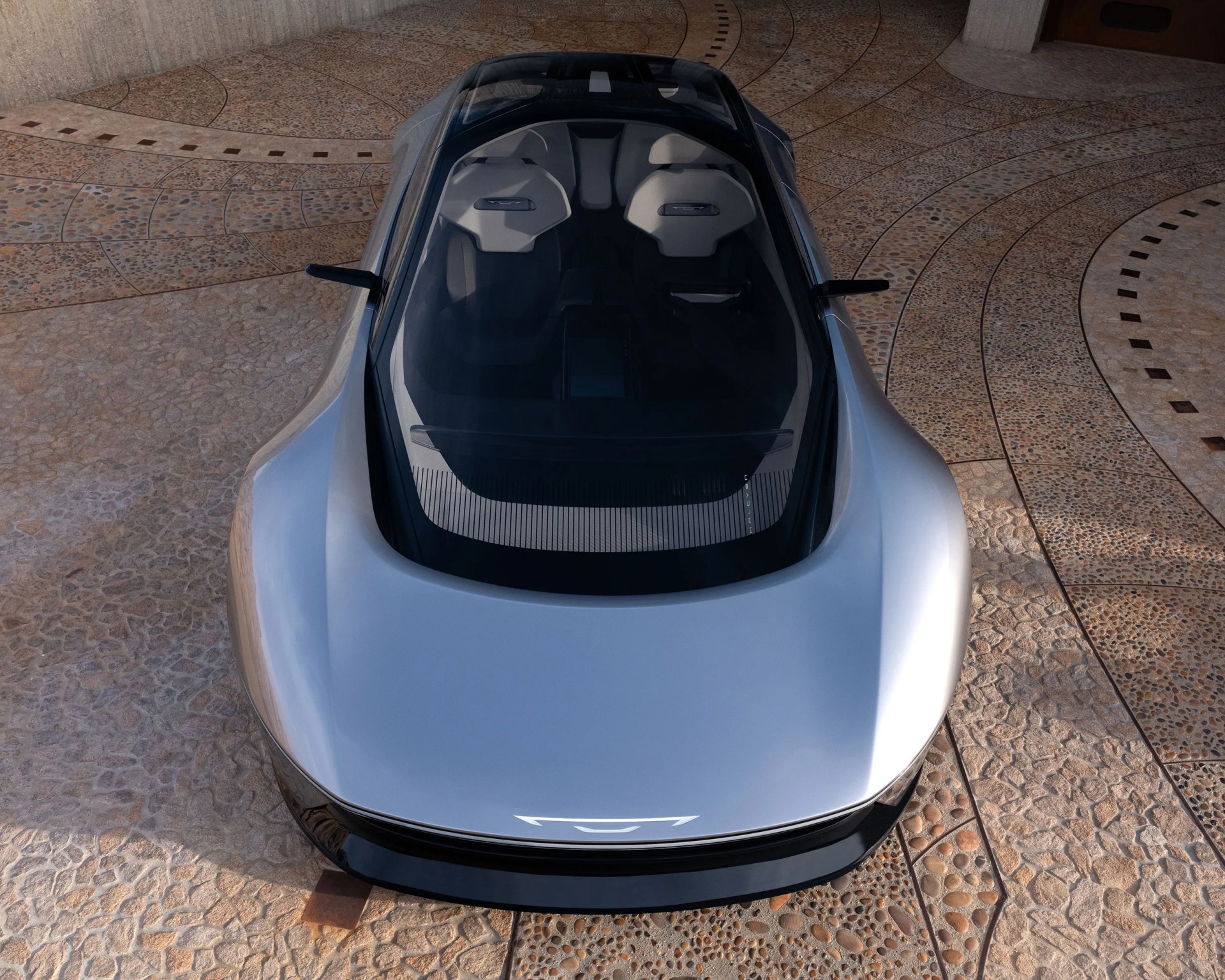 Silver sleek electric sports car with large glass roof and two white seats, parked on patterned stone pavement.