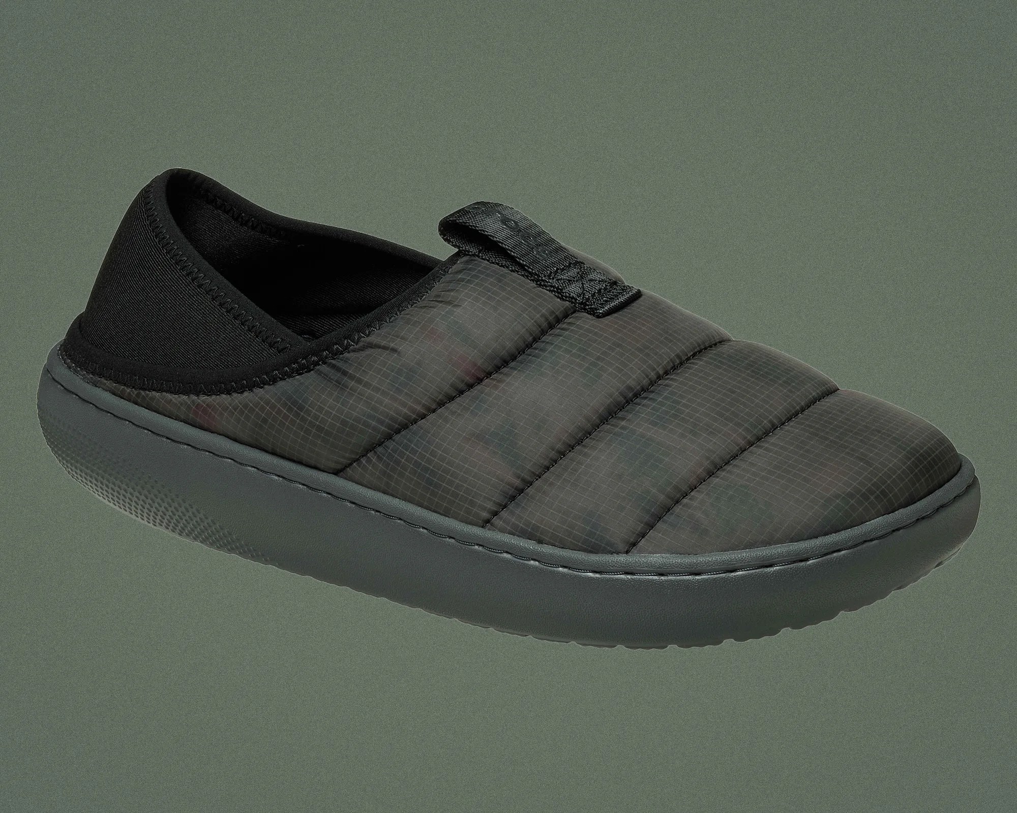 Black slip-on quilted slipper with padded upper and thick rubber sole on a green background.