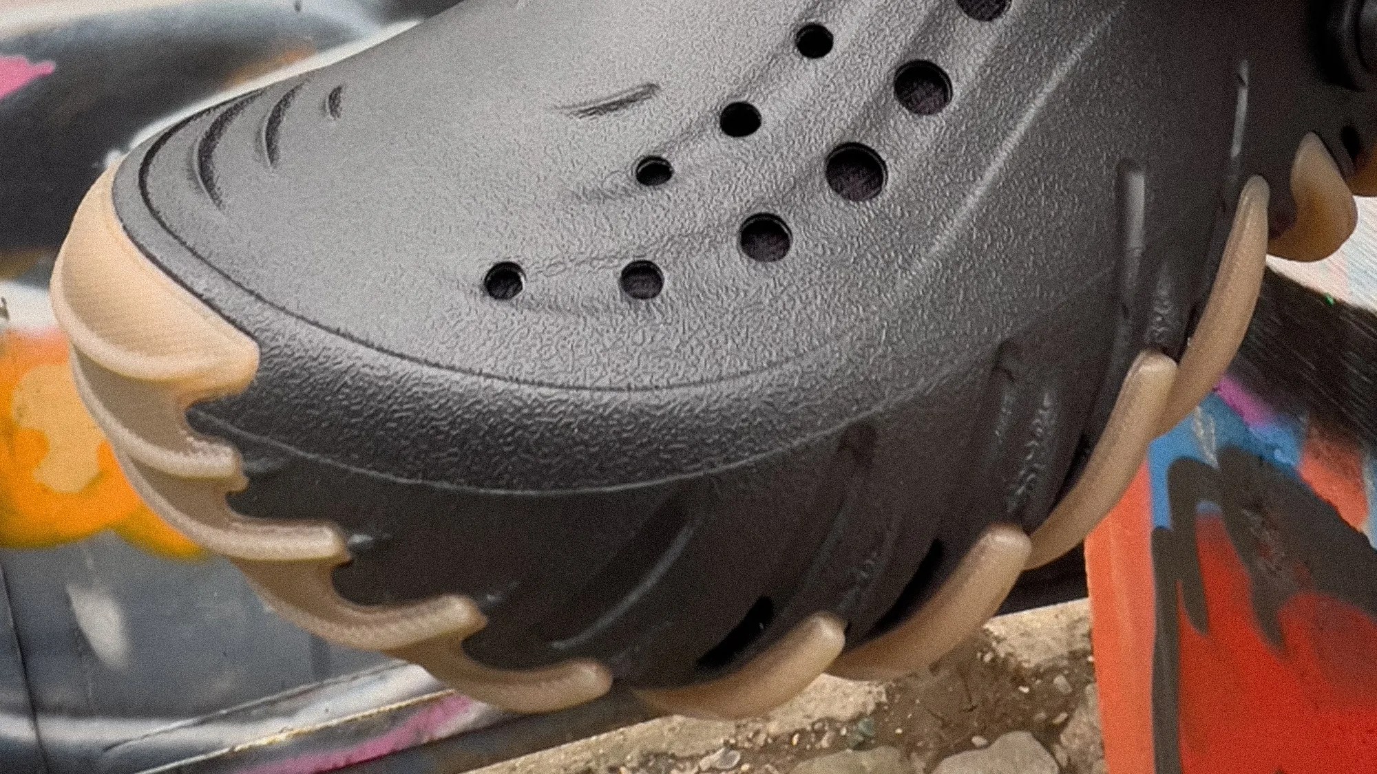 Close-up of a black rubber shoe with circular ventilation holes and a tan segmented sole against a graffiti background.