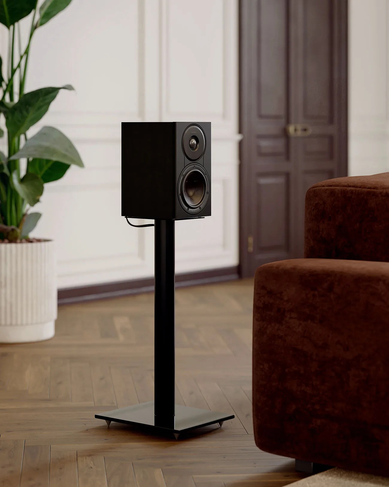 Black bookshelf speaker on a matching black metal stand on a wooden floor near a brown sofa and a potted plant.