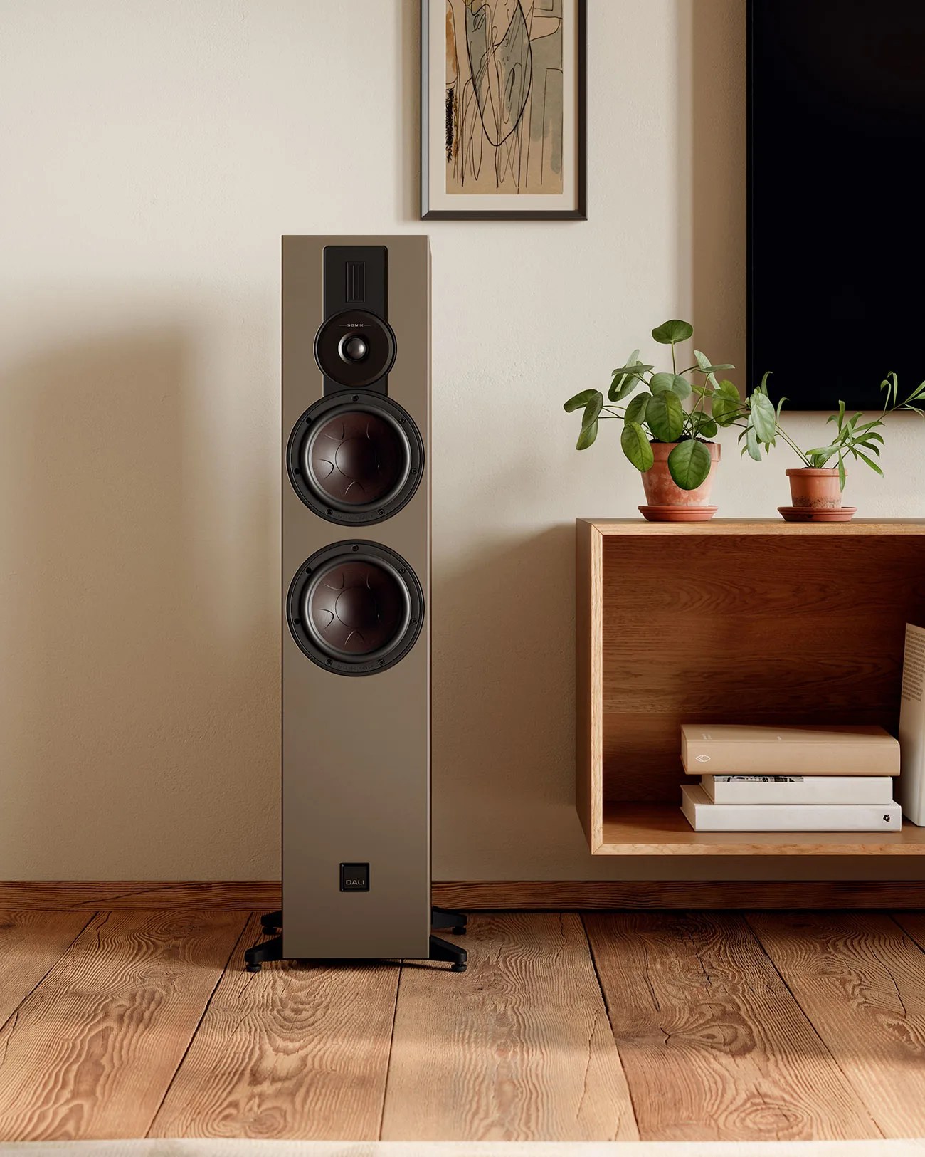 Tall beige floor-standing speaker with three drivers and black base on wooden floor next to a wooden shelf with plants and books.