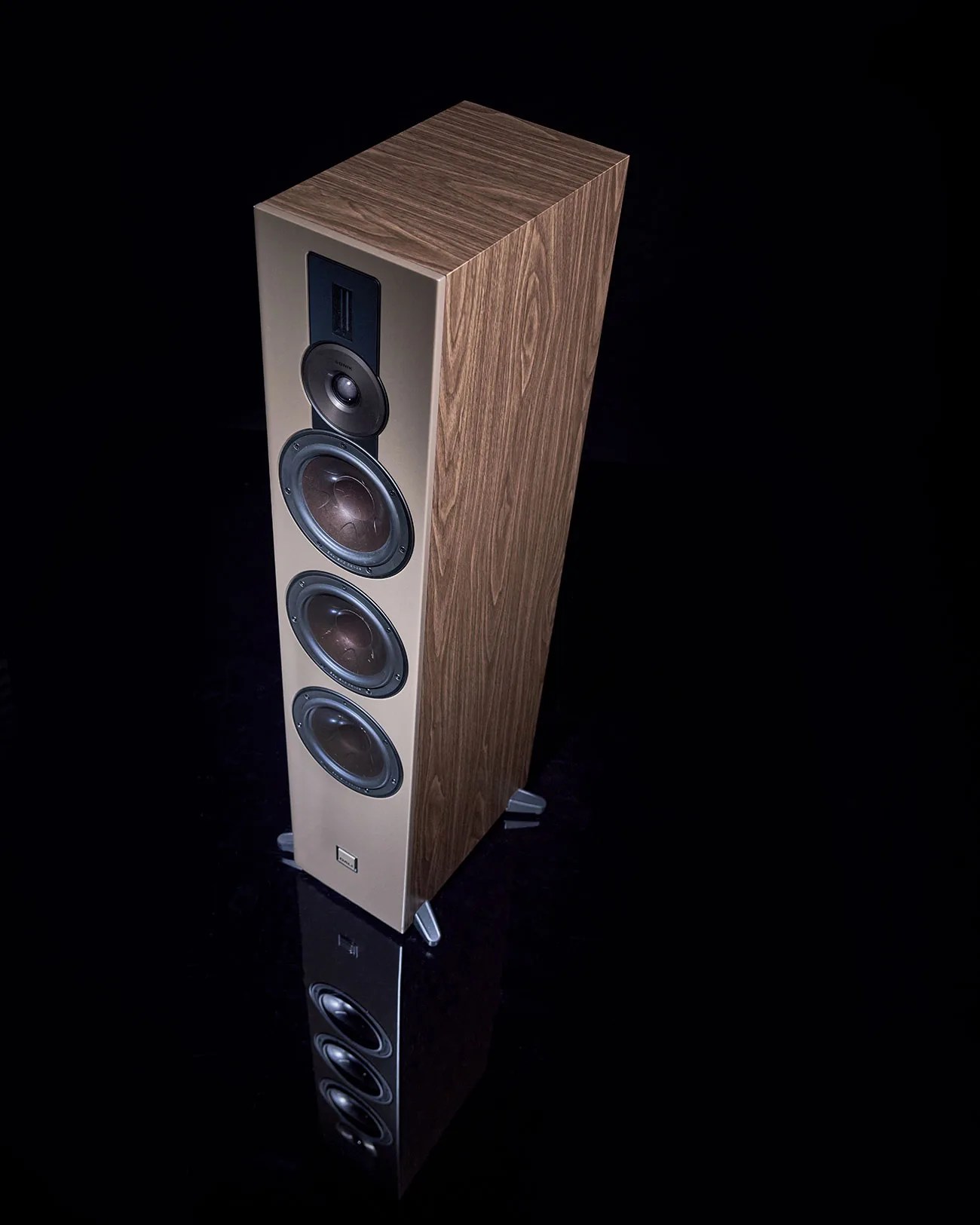 Tall floor-standing speaker with wood grain sides and four visible drivers on a beige front panel, reflected on a glossy black surface.
