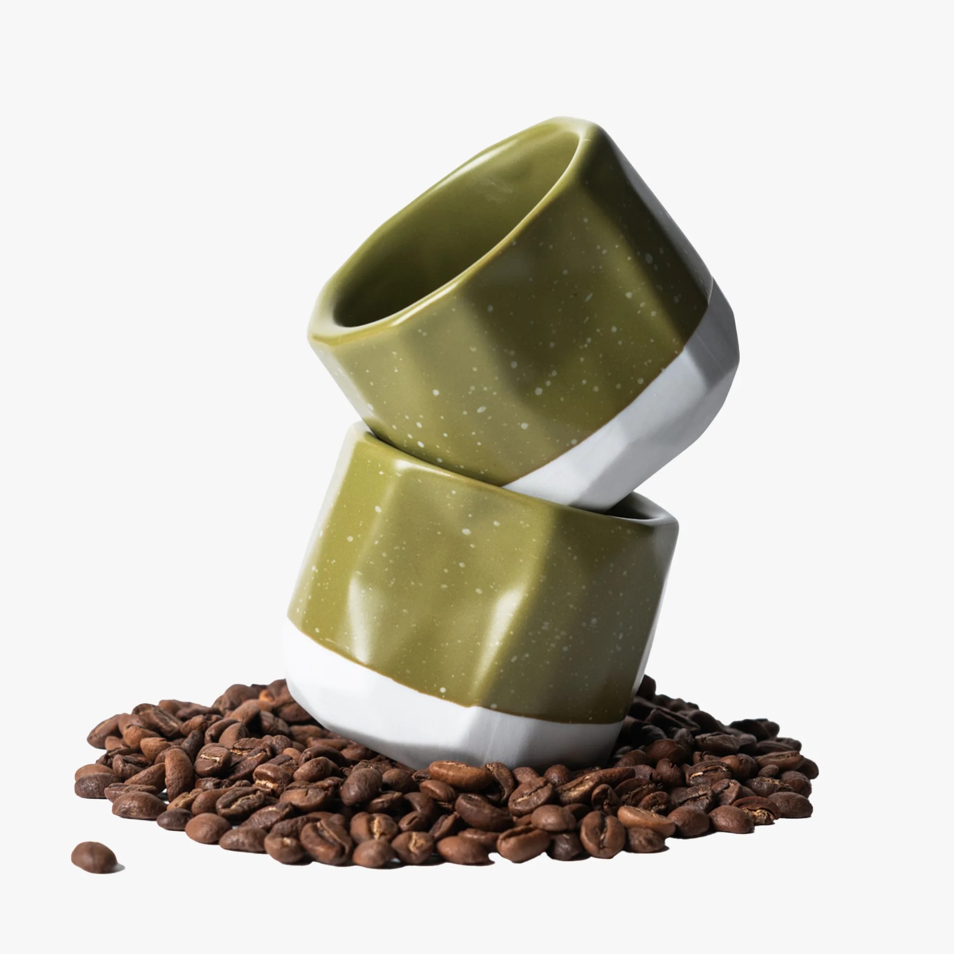 Two green and white ceramic cups stacked on a pile of coffee beans against a white background.