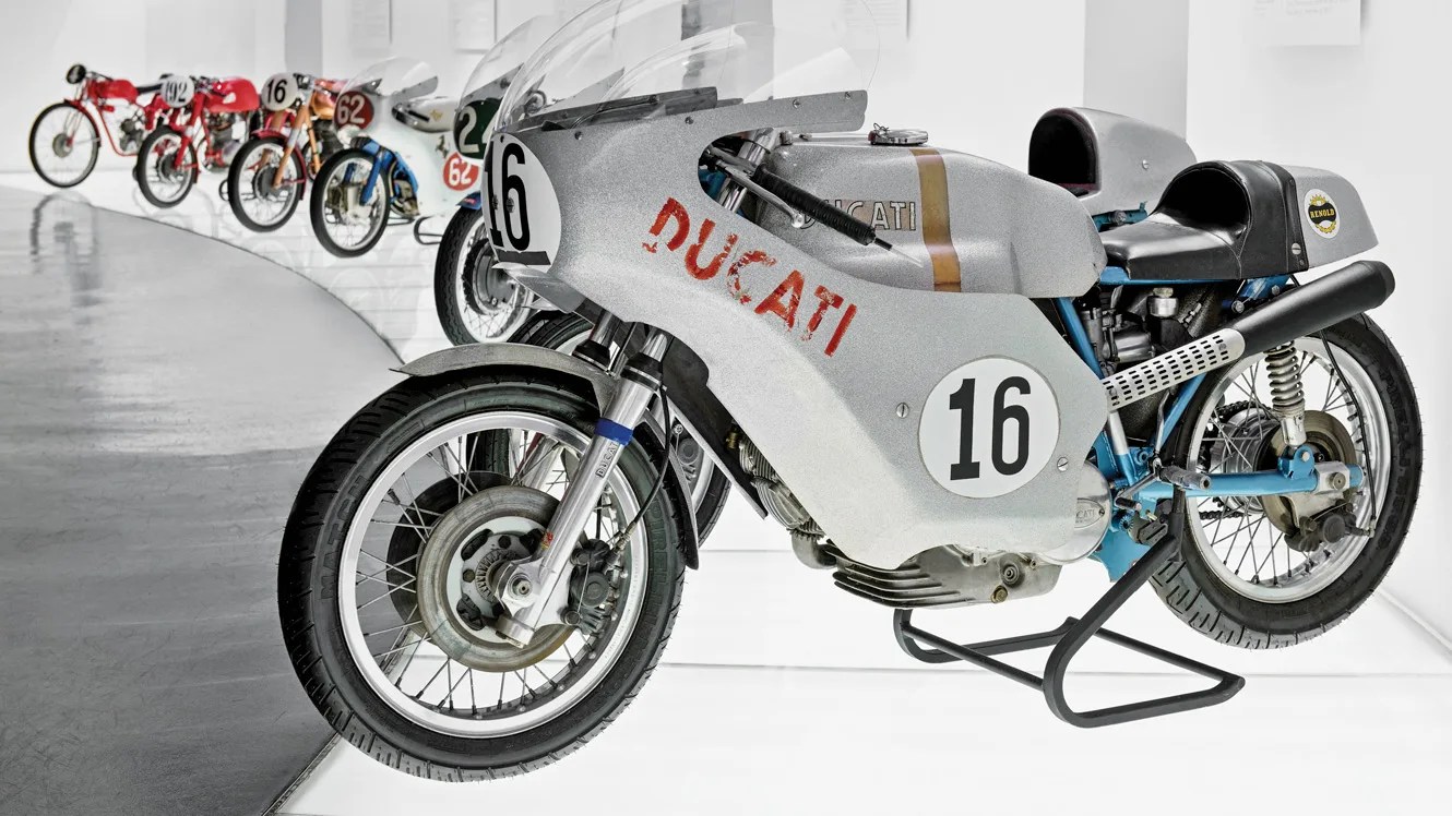 Row of vintage racing motorcycles with the closest one a silver Ducati numbered 16 with a black exhaust and blue frame.