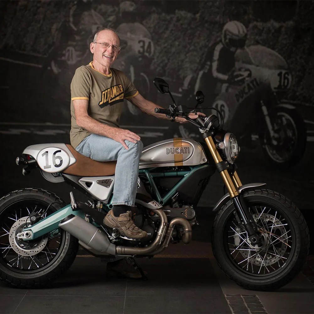 Man sitting on a silver Ducati motorcycle with brown seat and number 16 on the side panel.