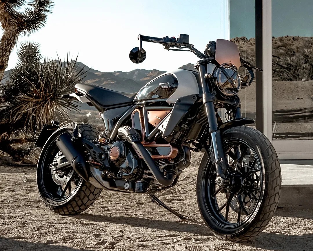 Black and copper scrambler motorcycle with knobby tires parked on sandy ground near desert plants.