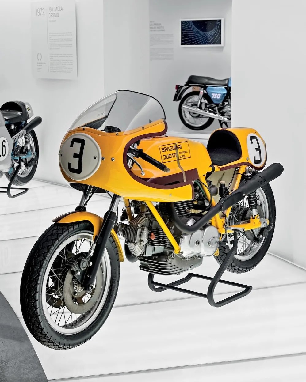 Yellow vintage Ducati racing motorcycle with number 3 on front and side, displayed on a stand in a museum.