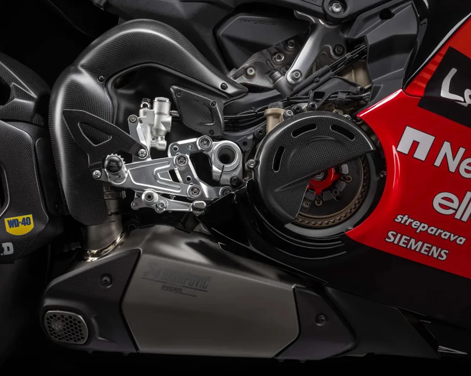 Close-up of a motorcycle's rear section showing a silver footpeg assembly, carbon fiber components, and a red fairing with sponsor logos.