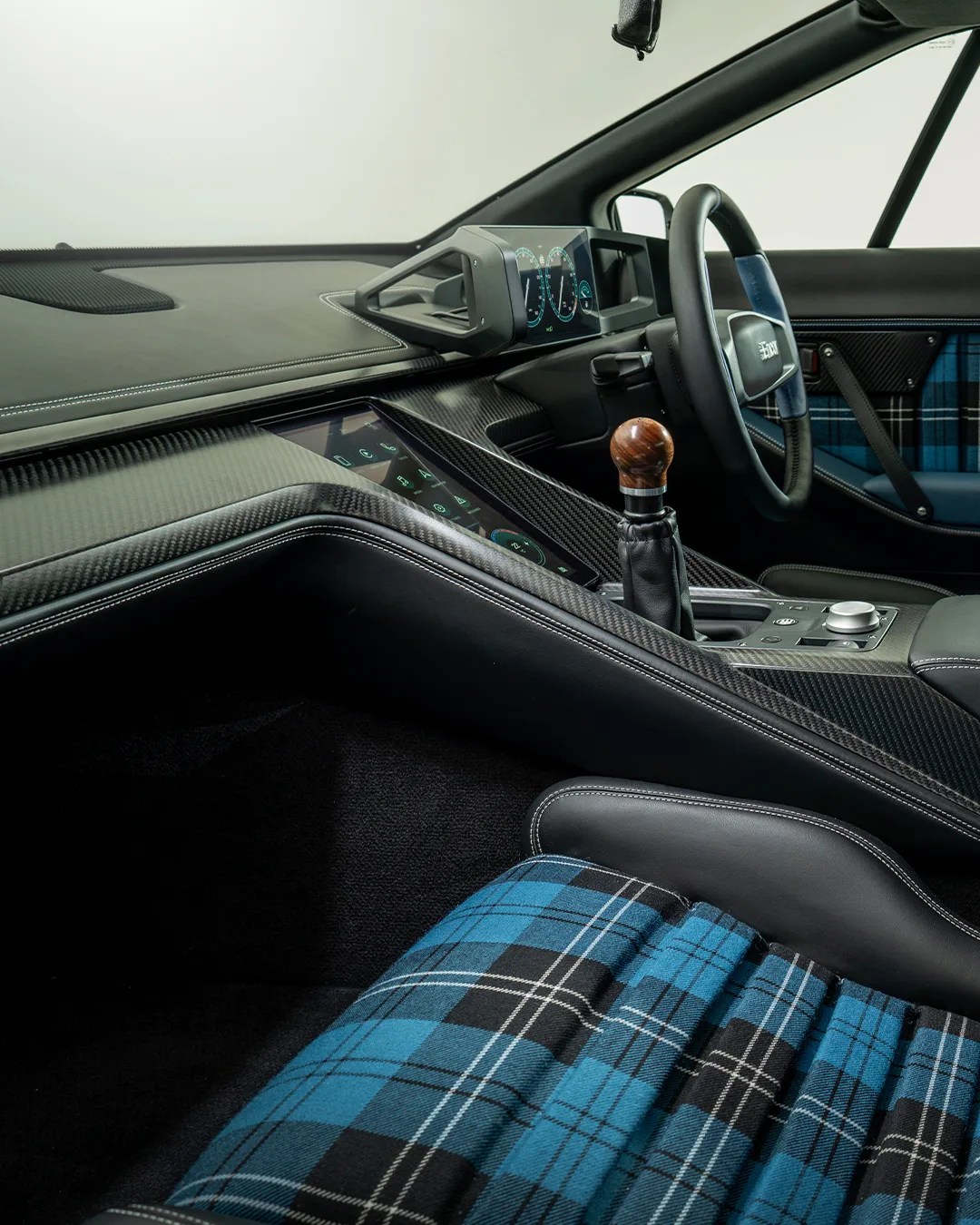 Car interior with carbon fiber dashboard, digital instrument cluster, wooden gear shift knob, and blue plaid fabric seats.