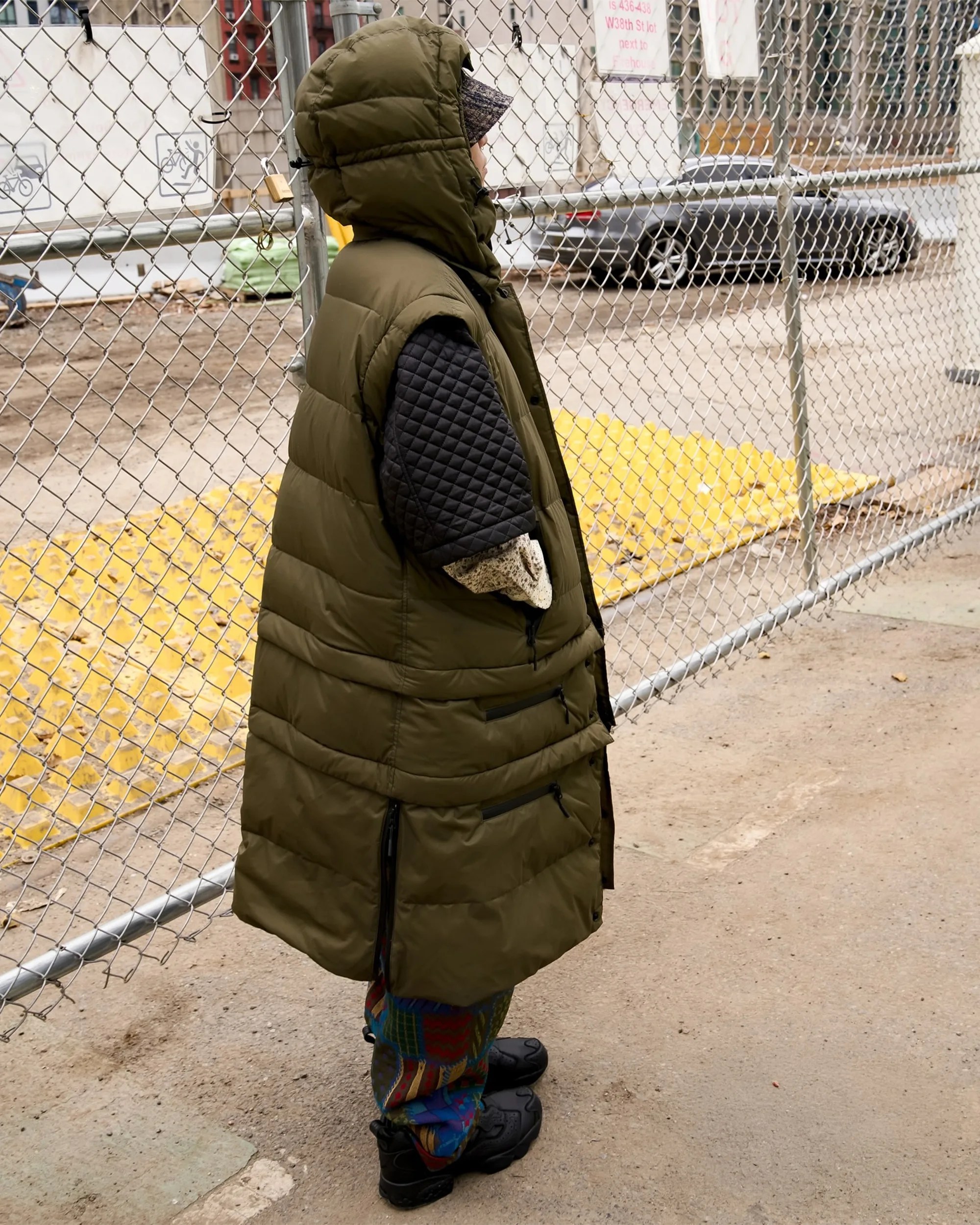 Person wearing an olive green long puffer vest with a hood, black quilted sleeves, and colorful pants standing by a chain-link fence.