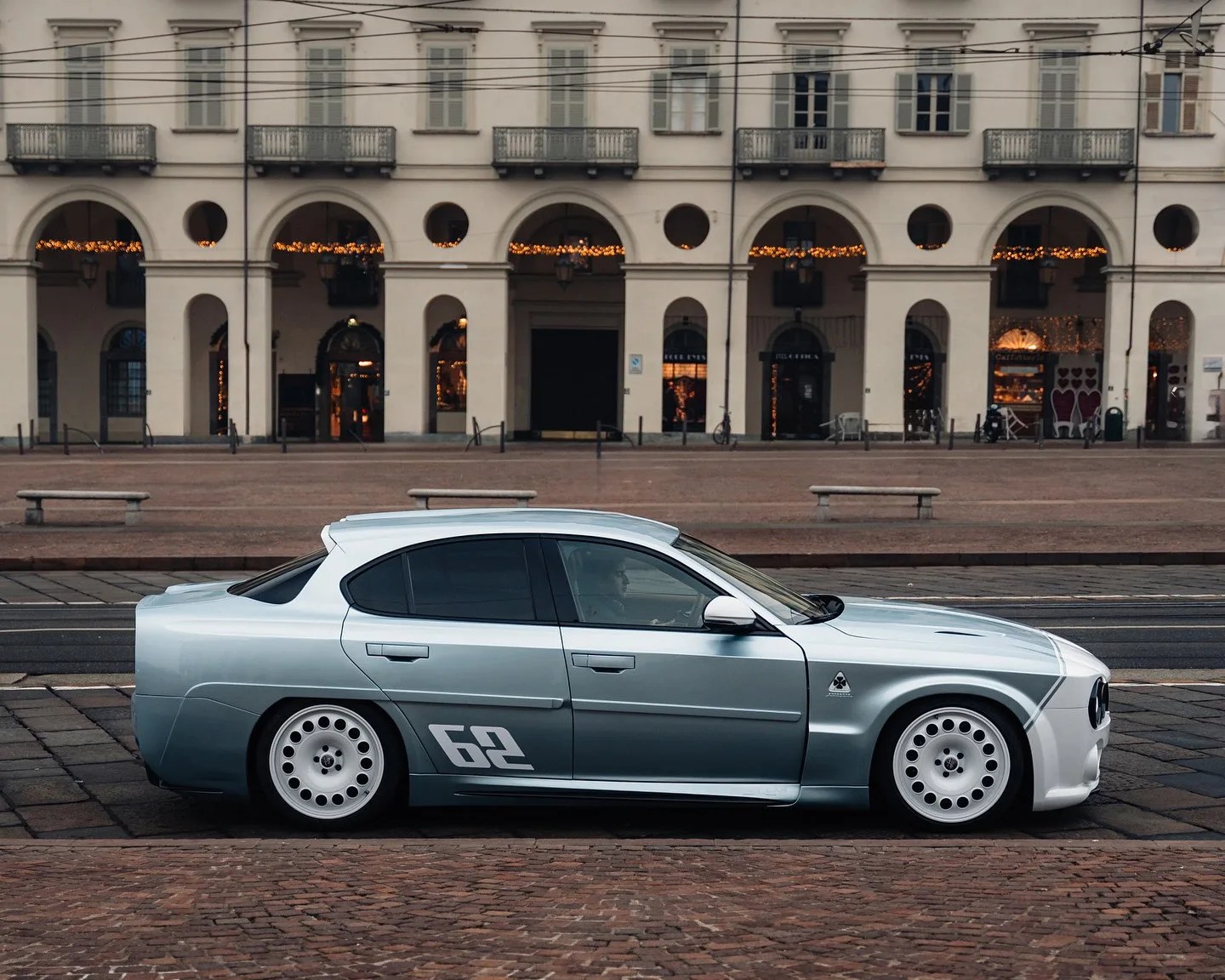 Silver and white retro-style sedan with number 62 decal parked on a cobblestone street in front of an arched building.