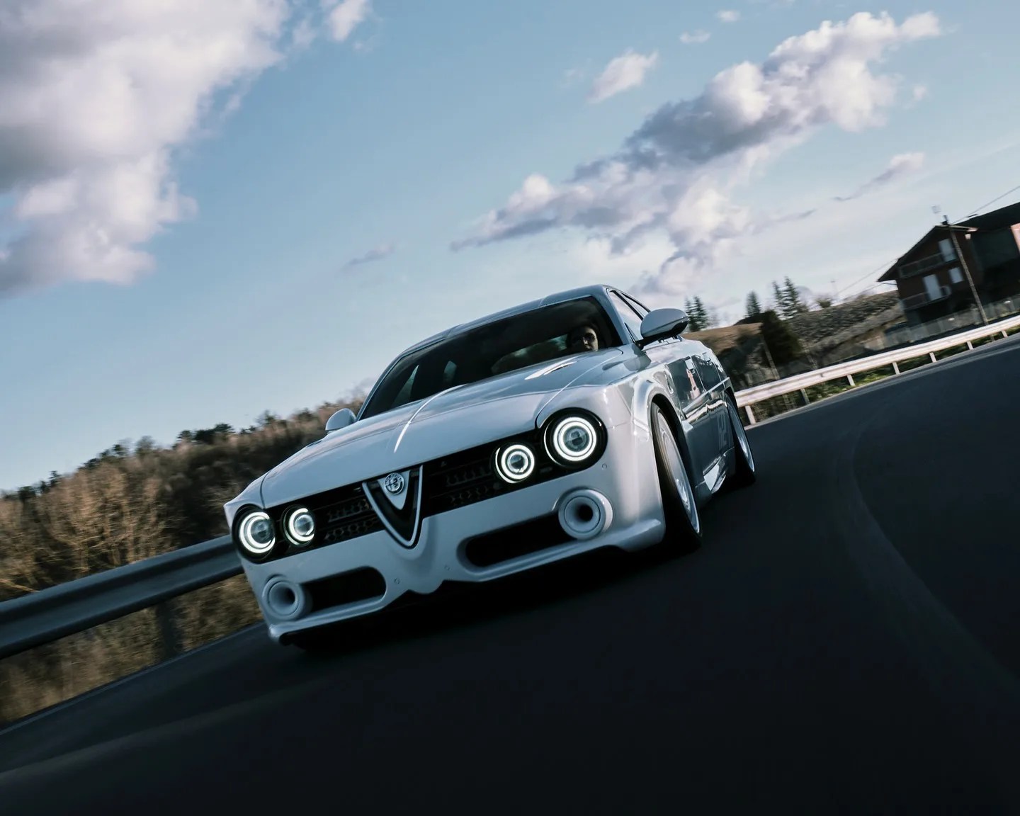 White Alfa Romeo car with circular LED headlights driving on a curved road with guardrails.