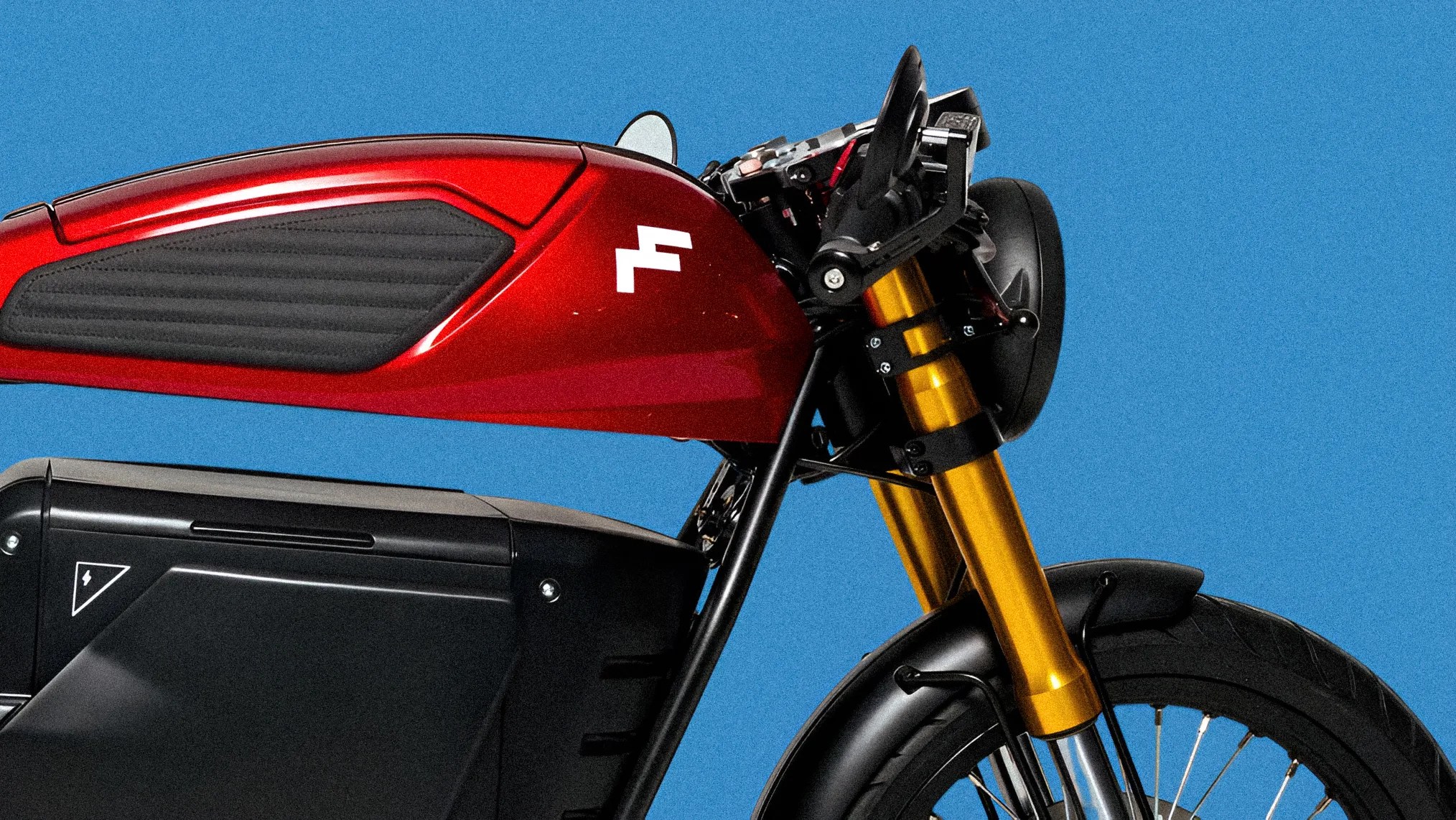Close-up of a red motorcycle fuel tank with a white logo, gold front forks, and black battery compartment against a blue background.