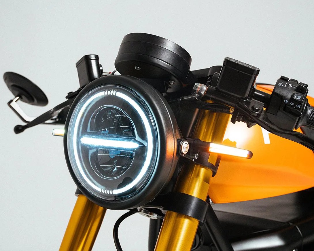 Round motorcycle headlight with LED halo and horizontal LED strip, mounted on gold front forks with an orange fuel tank in the background.