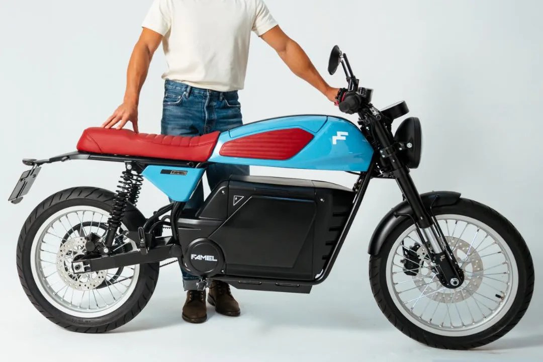 Blue and red Famel electric motorcycle with black battery casing and spoked wheels, held by a person in jeans and white shirt.