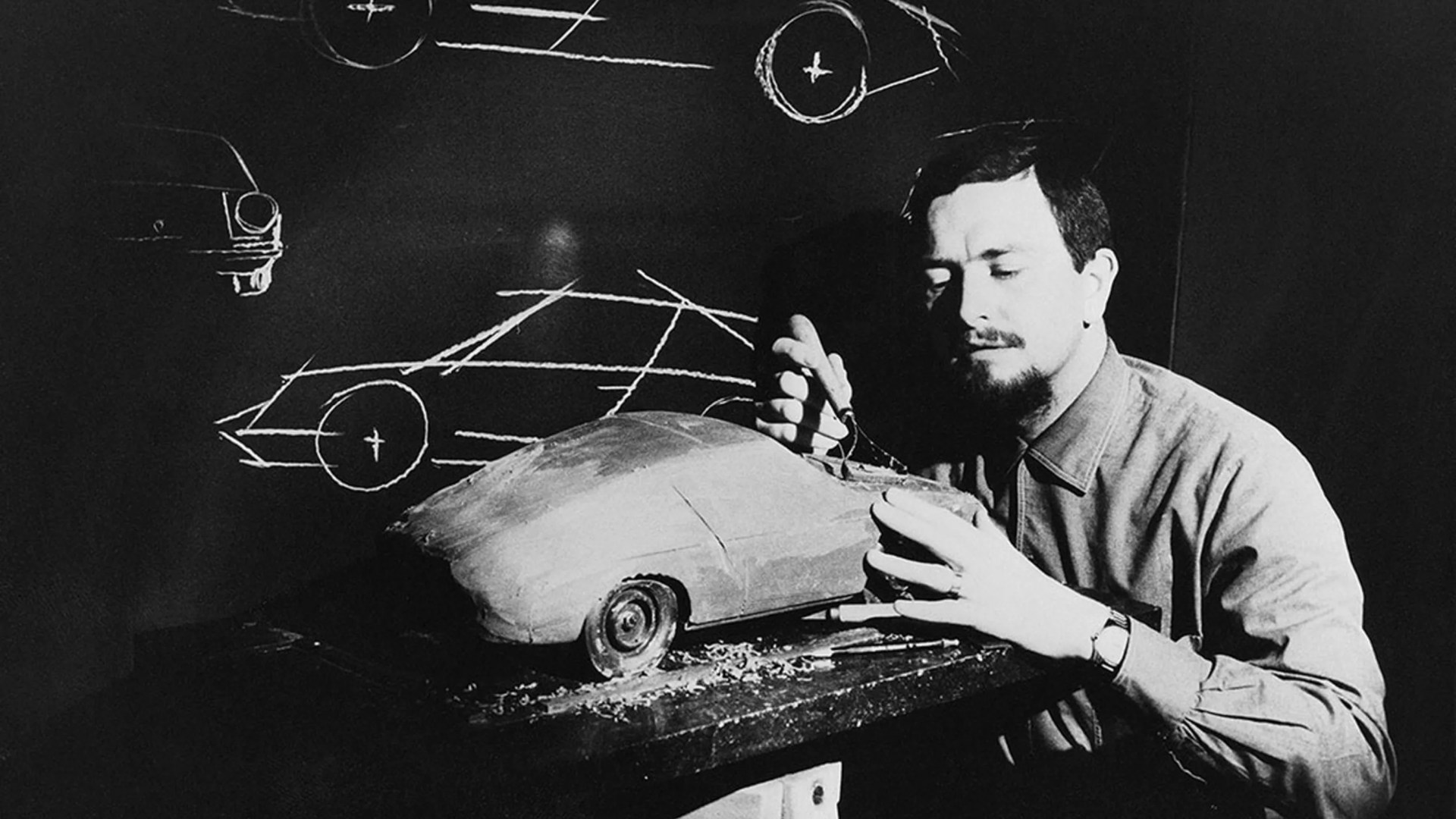 Man sculpting a clay model of a car on a table with chalk car sketches on a blackboard behind him.