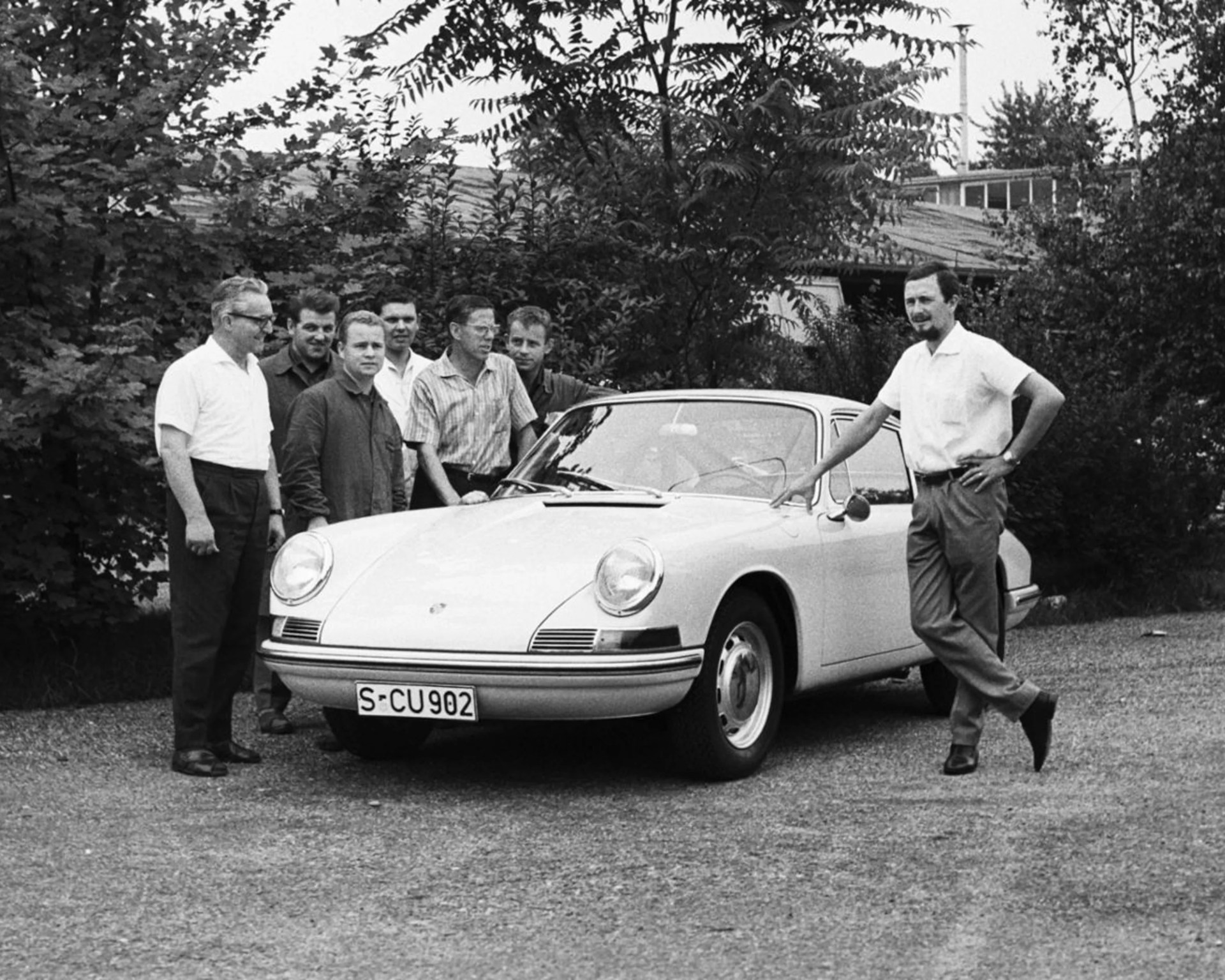 Black and white photo of six men standing around a classic Porsche 911 with license plate S-CU 902 outdoors.
