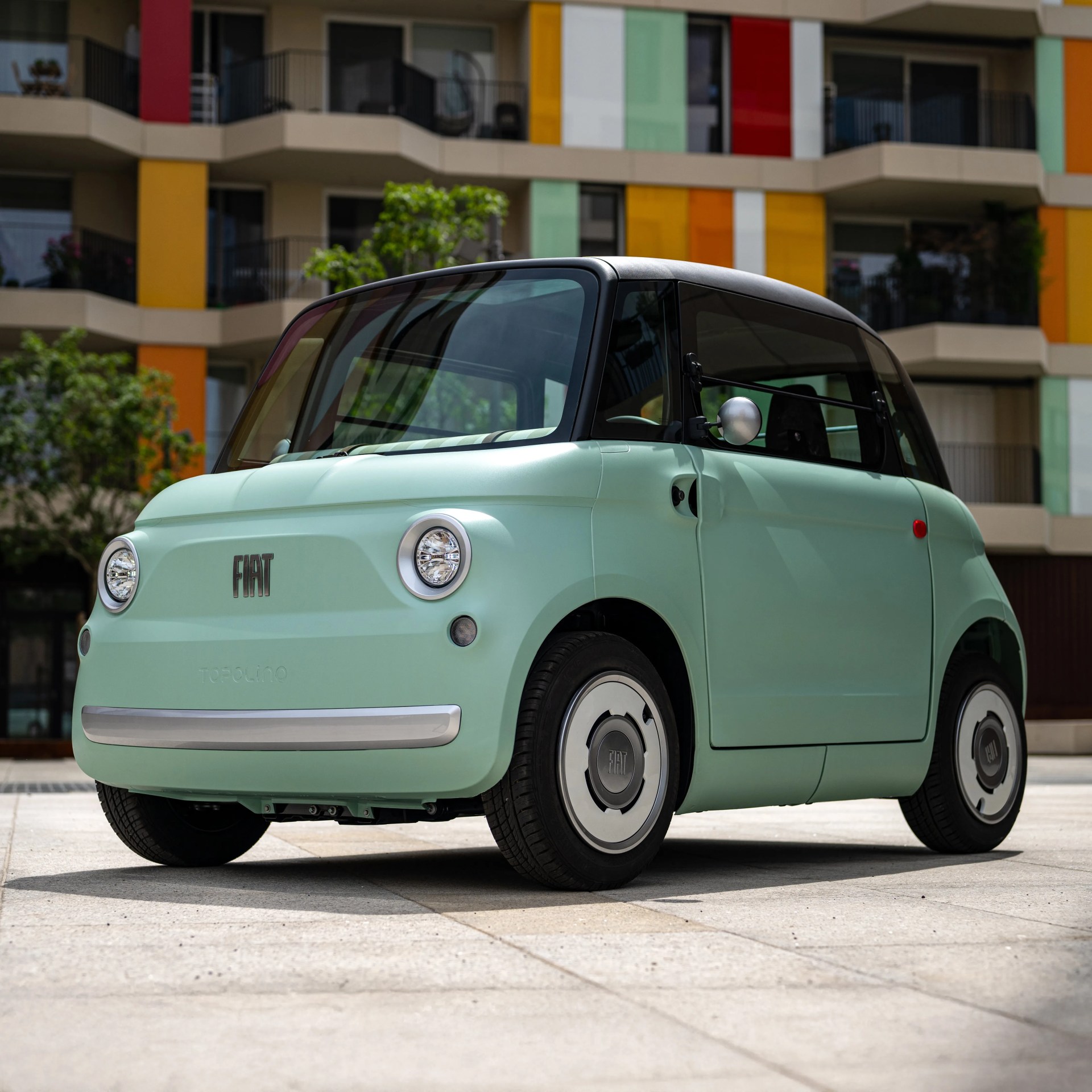 Mint green Fiat Topolino electric car with black roof parked in front of a colorful apartment building.