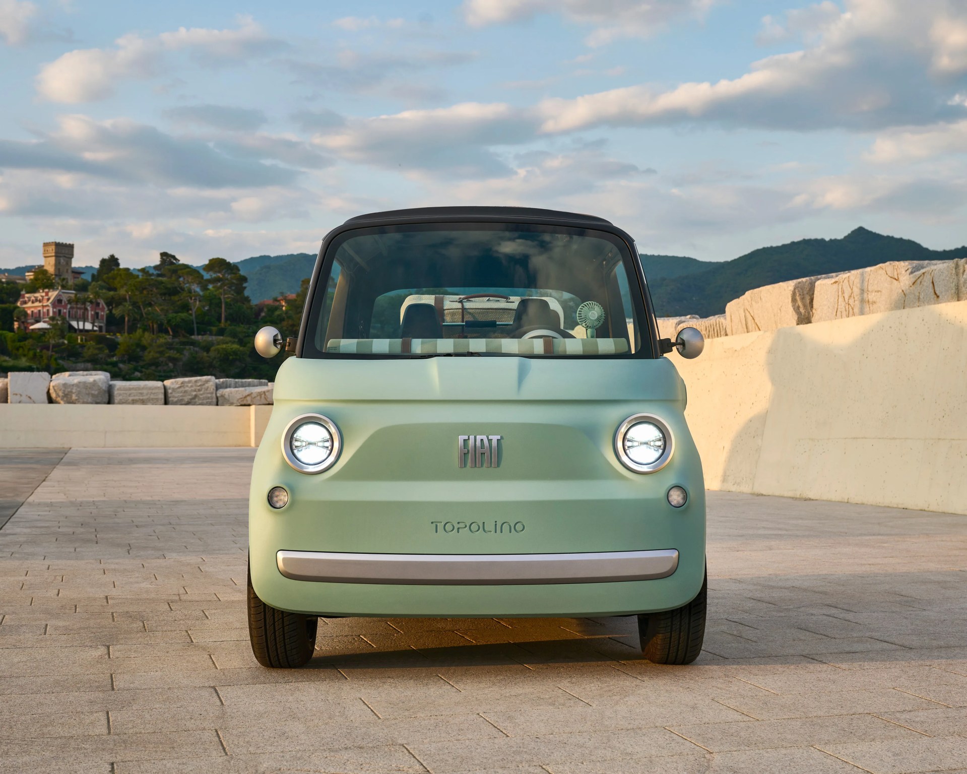 Front view of a light green Fiat Topolino compact car with round headlights and black roof parked on a stone pavement.