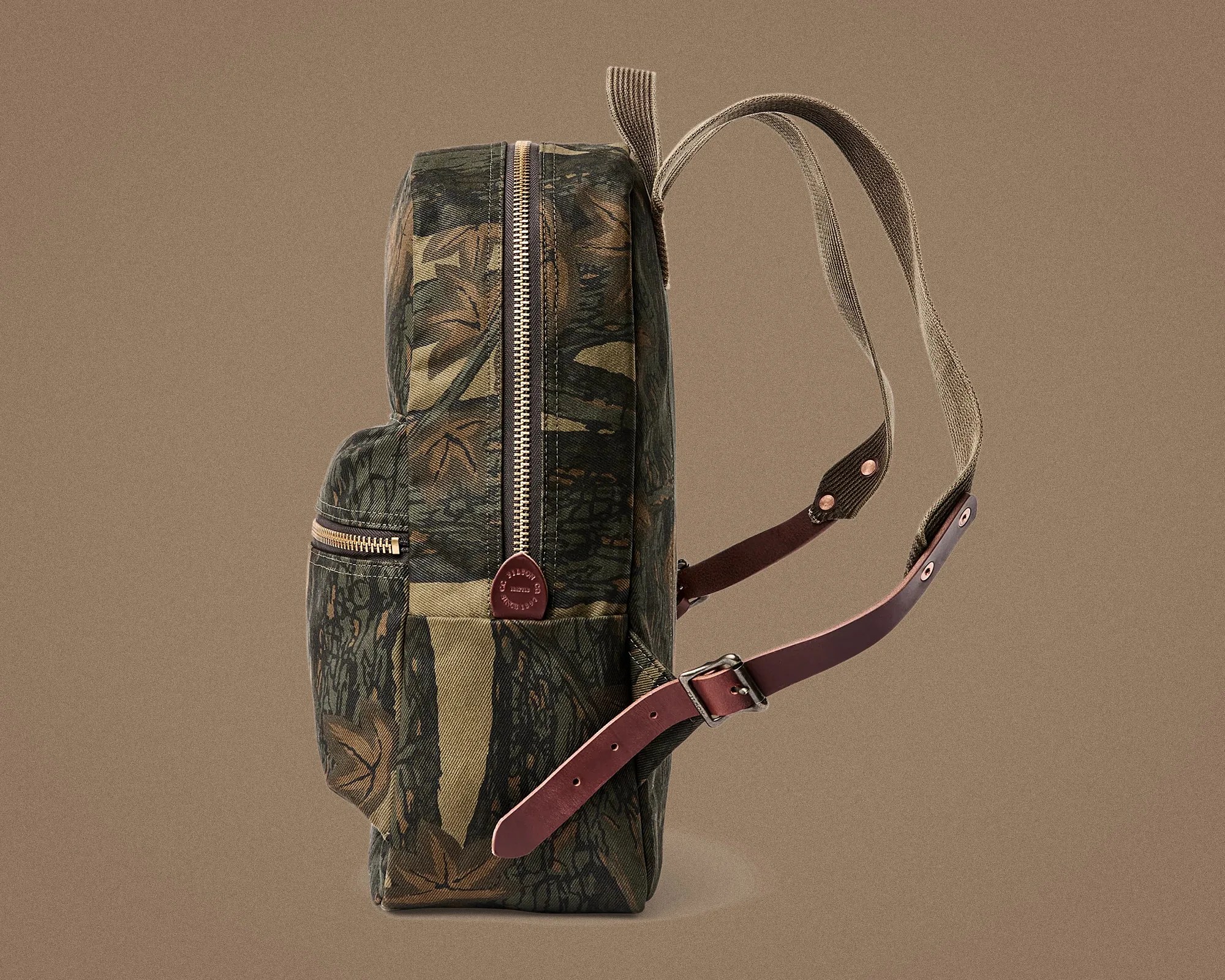 Side view of a camouflage-patterned backpack with tan fabric straps and brown leather adjustable buckles.