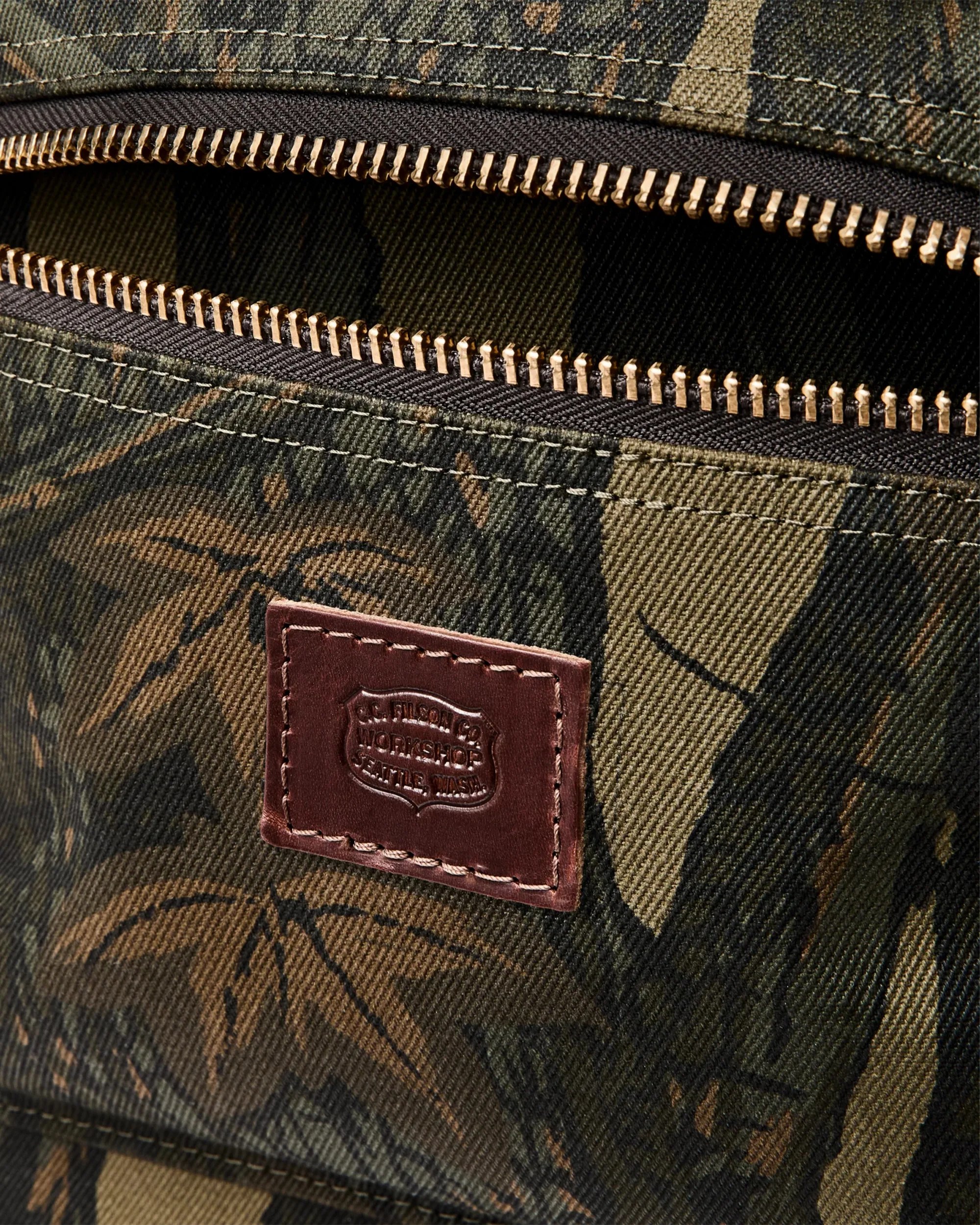 Close-up of a camouflage fabric bag with a brass zipper and a brown leather patch embossed with "J.C. Filson & Co Workshop Seattle Wash.