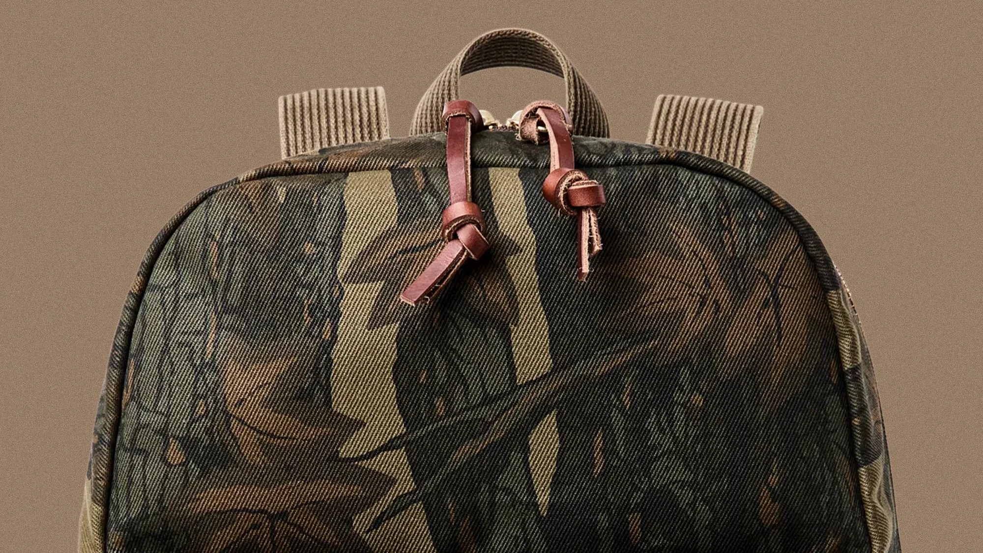 Backpack with camouflage fabric, leather zipper pulls, and ribbed fabric straps against a beige background.