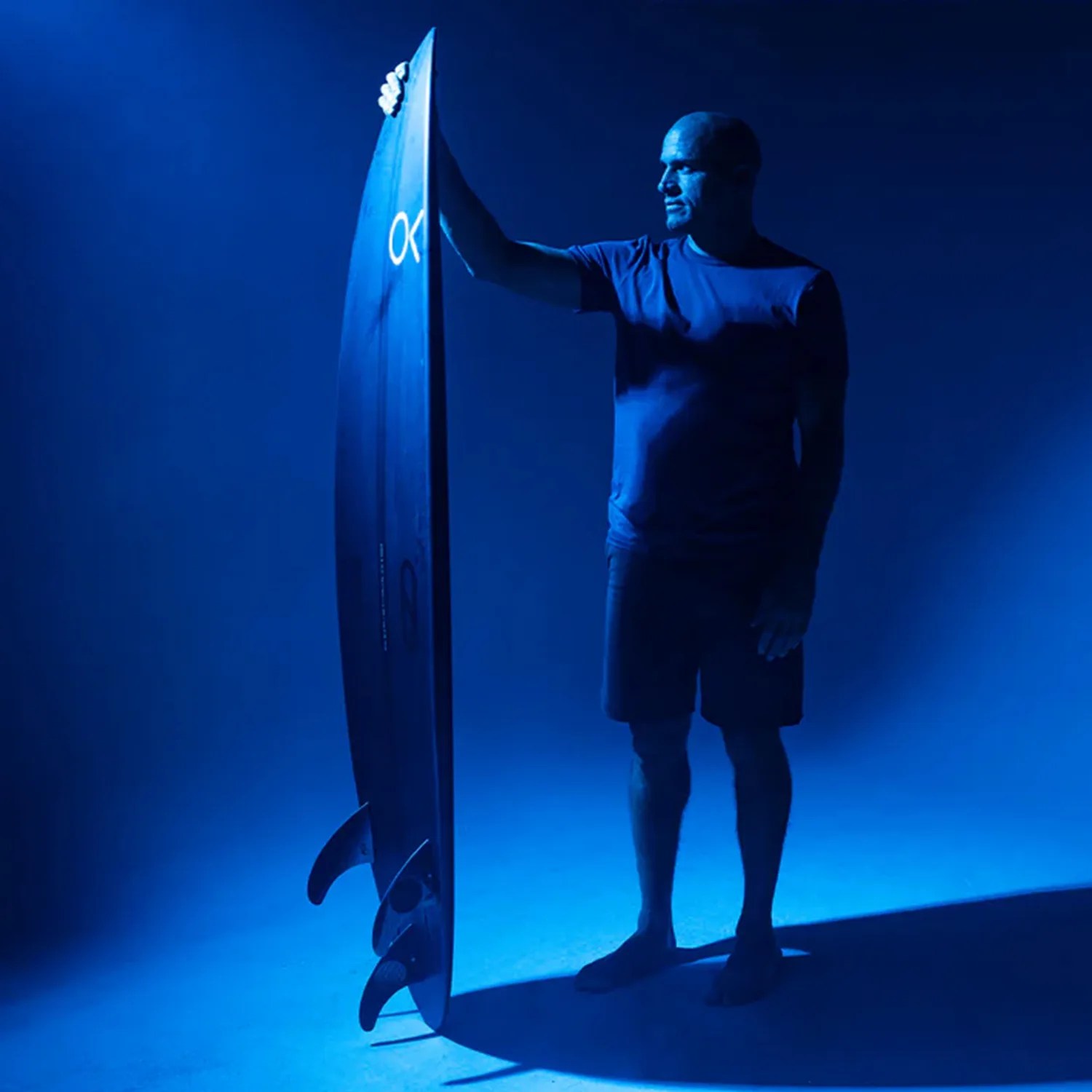 Man in a blue shirt and shorts holding a black surfboard with three fins in blue lighting.