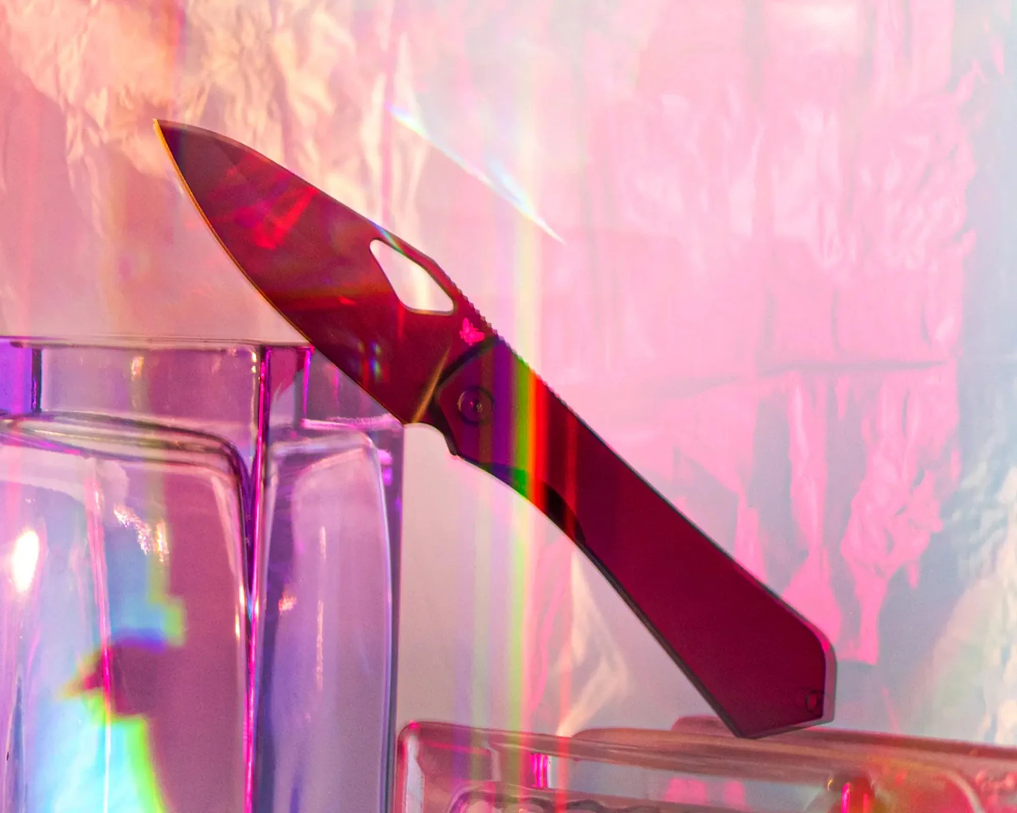 Red folding knife with a partially serrated blade resting on clear glass blocks with pink and purple lighting.