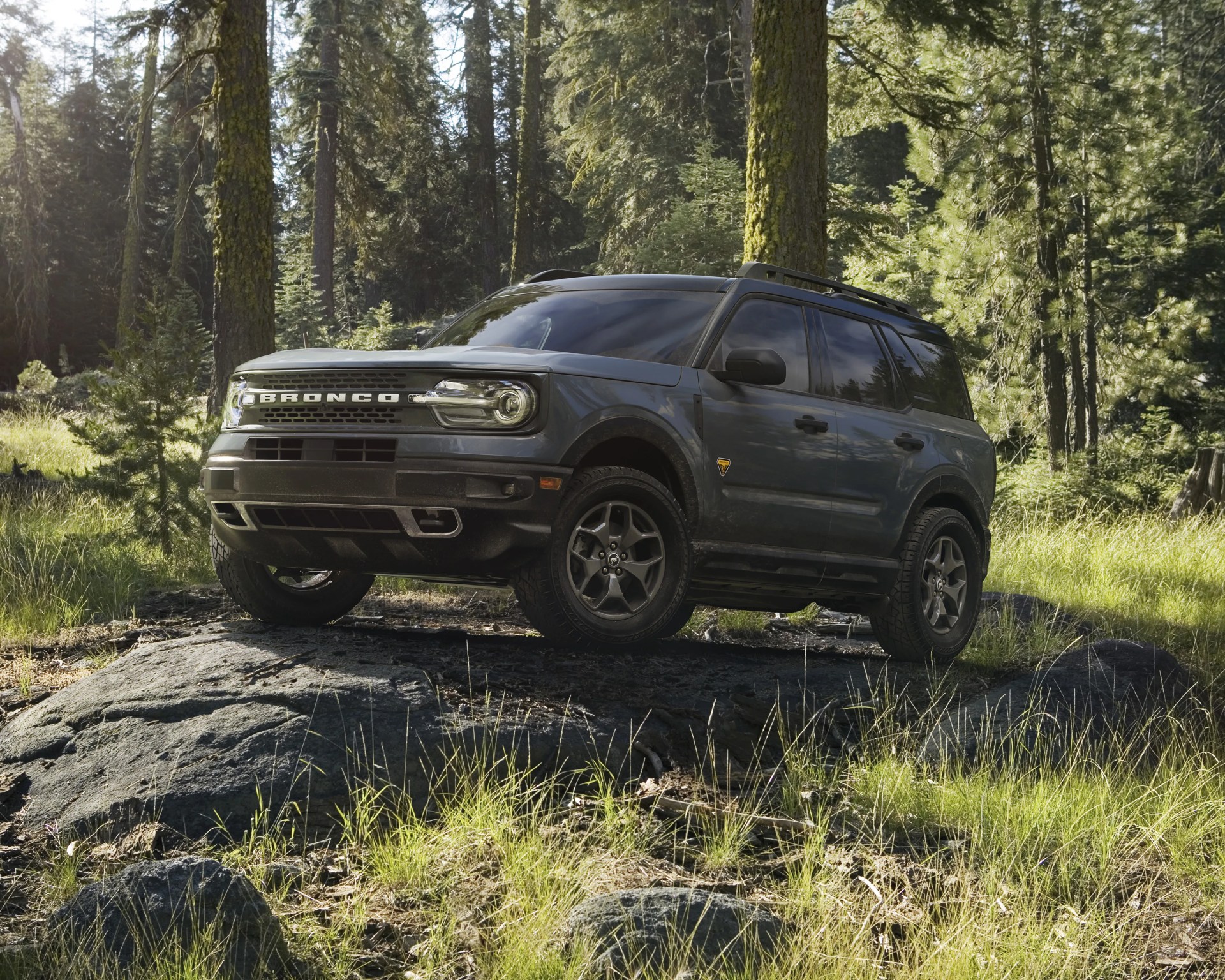 Dark gray Ford Bronco SUV parked on a large rock in a sunlit forest clearing.