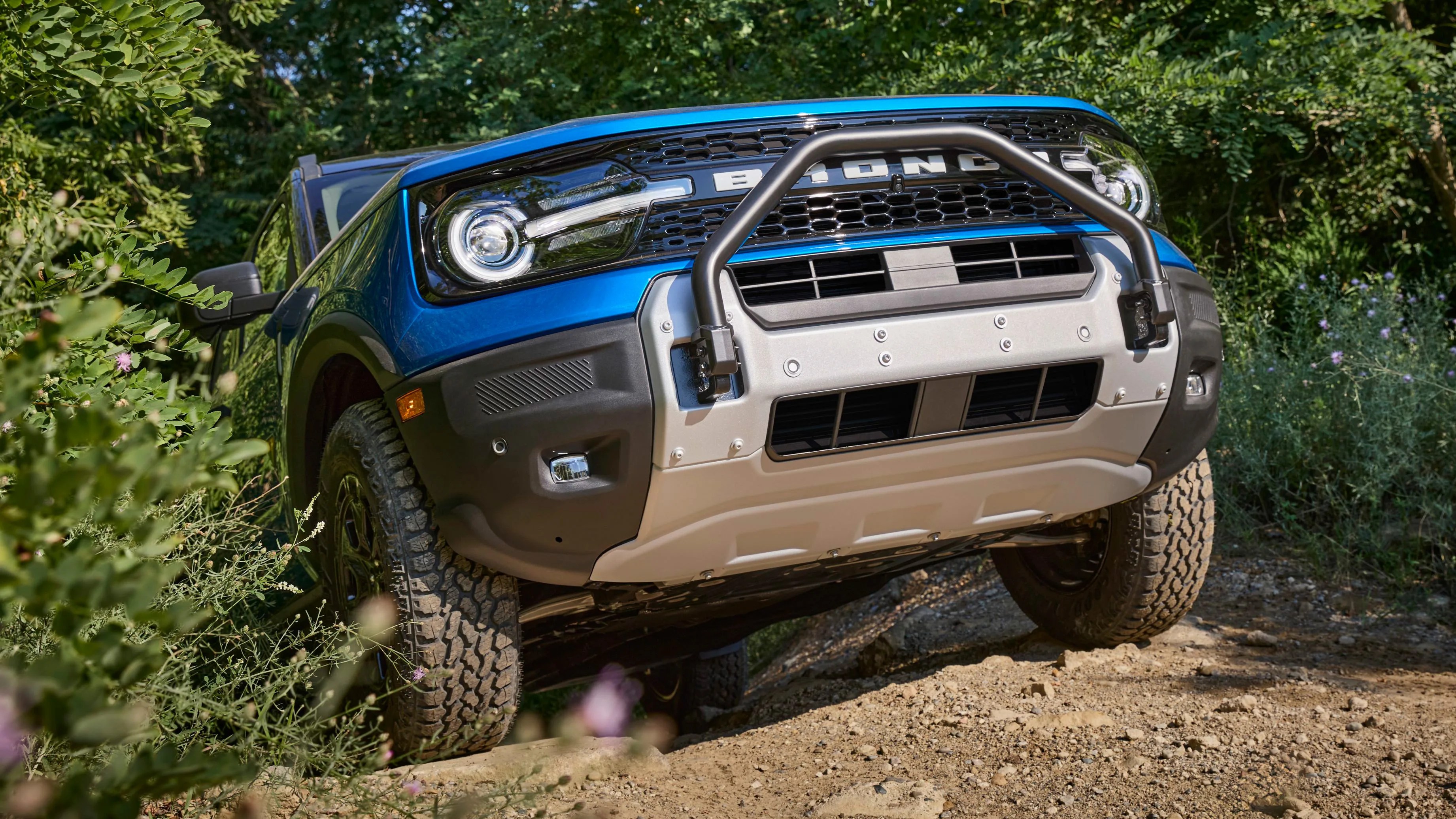 Blue Ford Bronco with off-road tires and front bull bar driving on a dirt trail surrounded by greenery.