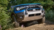 Blue Ford Bronco with off-road tires and front bull bar driving on a dirt trail surrounded by greenery.